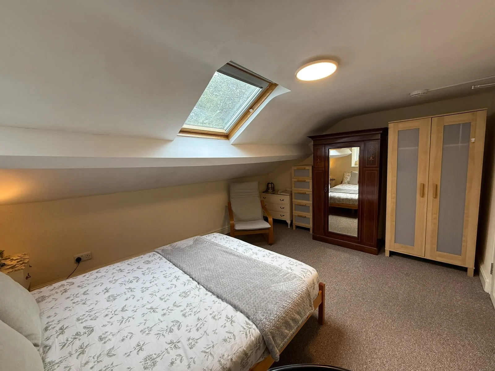 A cozy bedroom with a sloped ceiling and skylight window. There is a bed with floral bedding, a wooden chair, a white dresser, and two wardrobes, one with a mirror. A lamp on the wall provides warm lighting.