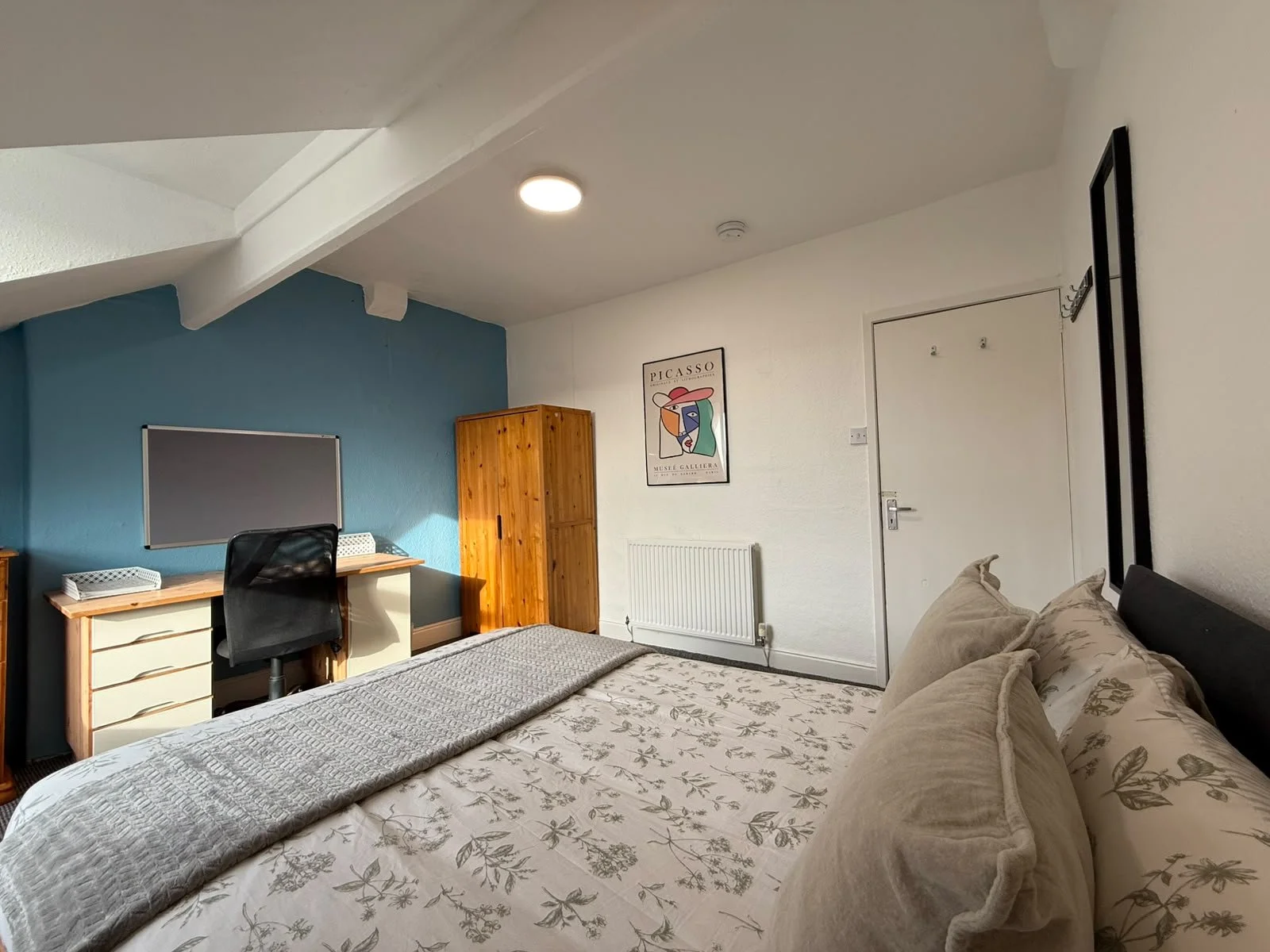 A bedroom with a bed featuring beige floral bedding and pillows, a desk with a black office chair, a wooden wardrobe, a radiator, and a wall decoration of Picasso artwork.