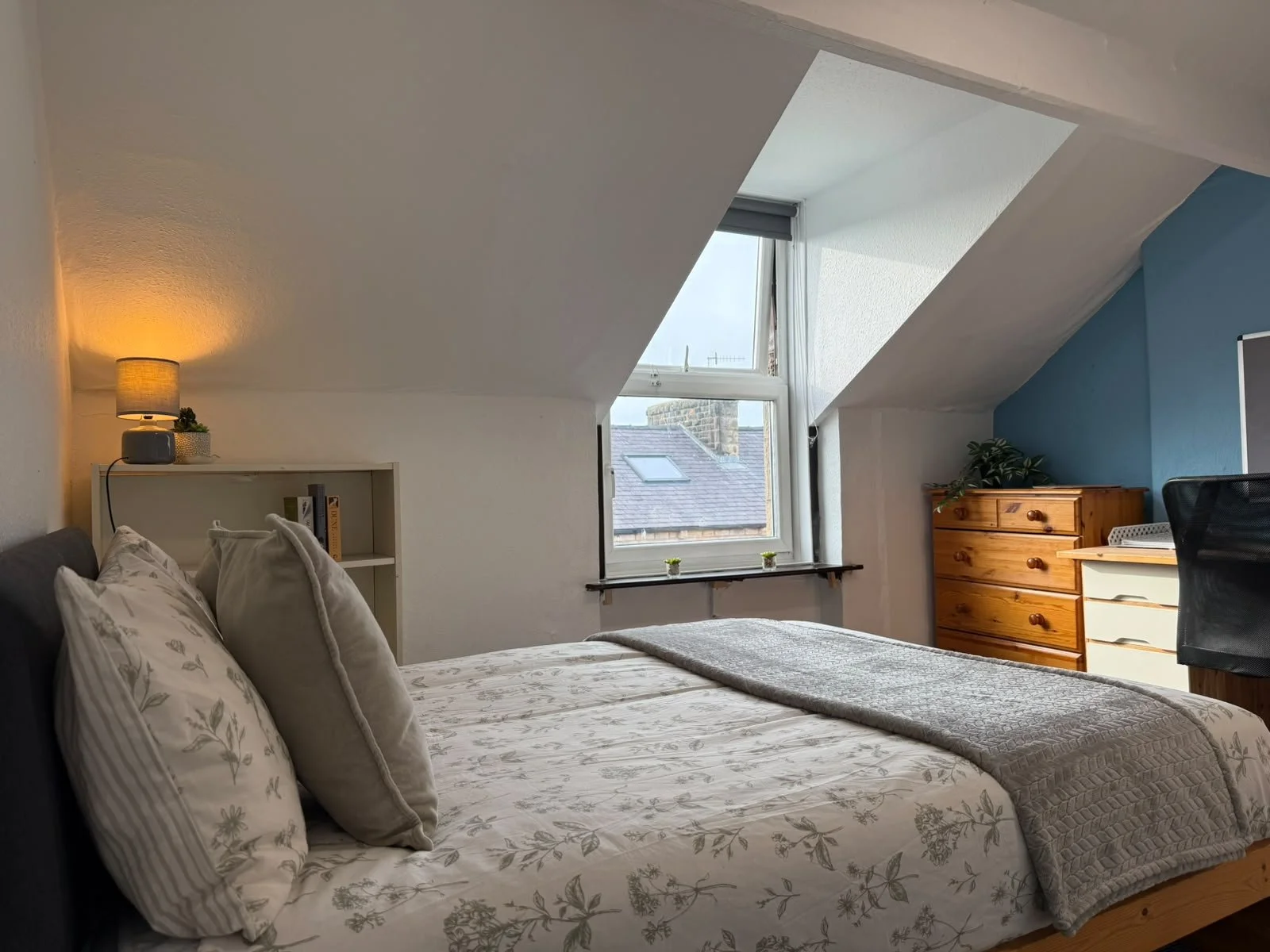A bedroom with sloped ceiling, a bed with floral bedding and pillows, a window with a view of rooftops, a small desk, a wooden dresser, a lamp, and a black office chair.