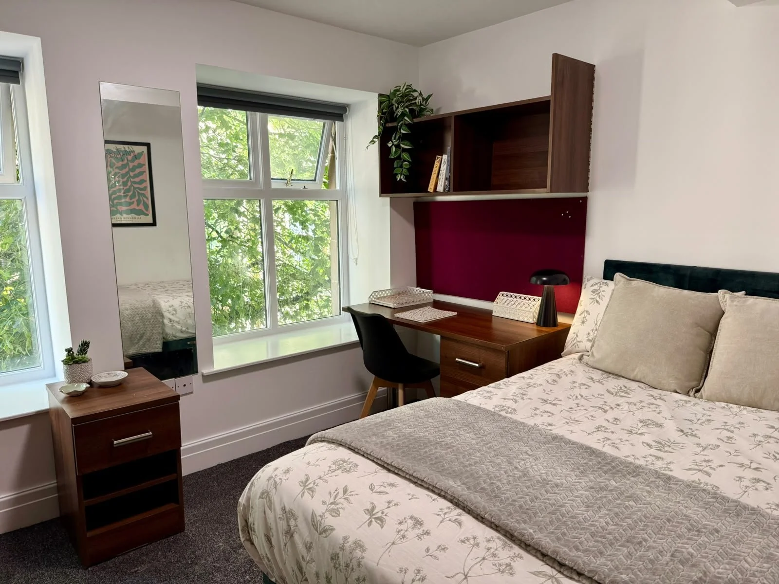 A cozy bedroom with a large window showing green leaves outside. There is a bed with floral bedding and pillows, a small wooden nightstand with a plant and dishes, a mirror on the wall, and a study area with a desk, chair, black lamp, and wooden shel