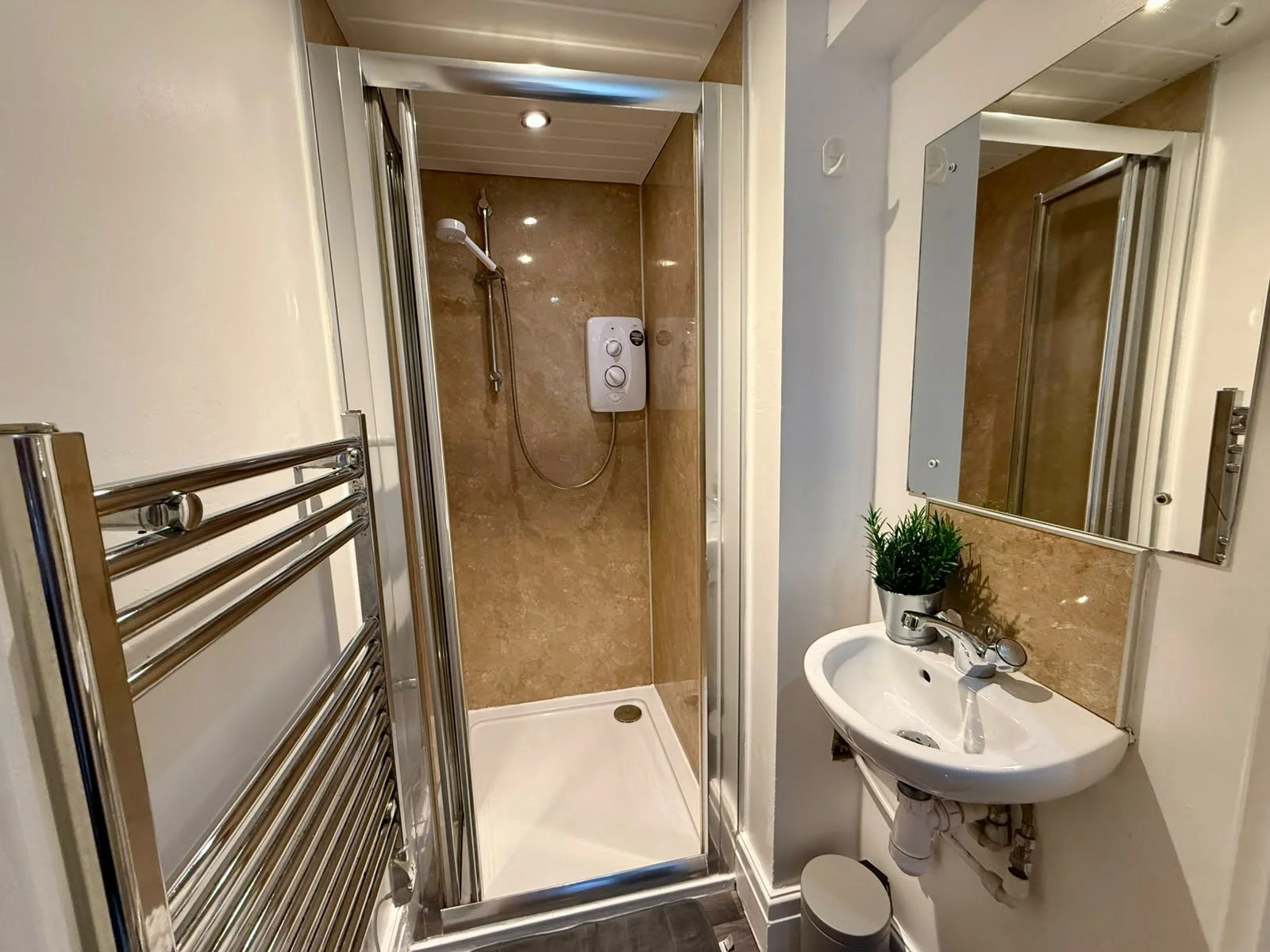 A compact bathroom featuring a small shower with beige marble walls and a white shower base, a wall-mounted mirror above a small white sink with a plant on it, a towel radiator on the left, and a small trash bin underneath the sink.