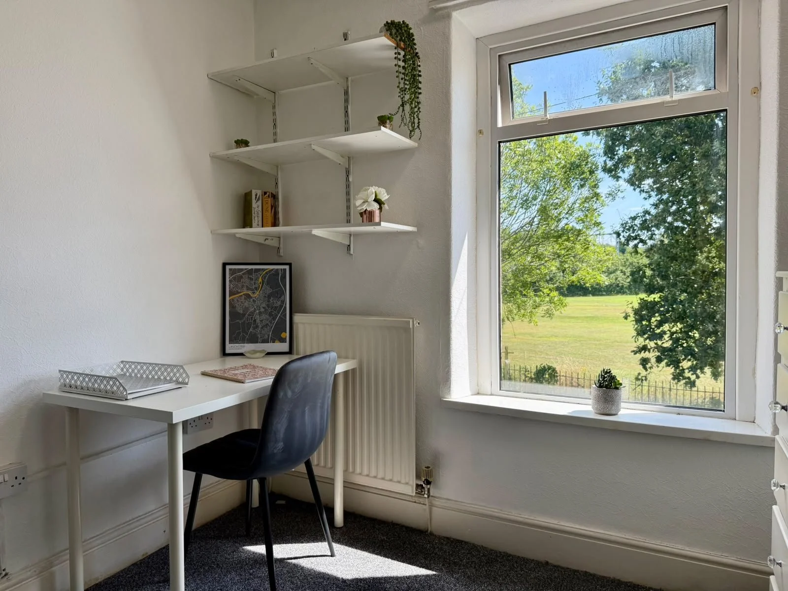 A small workspace corner with a white desk, black chair, white wall-mounted shelves with plants, a framed map, and a window showing greenery outside.