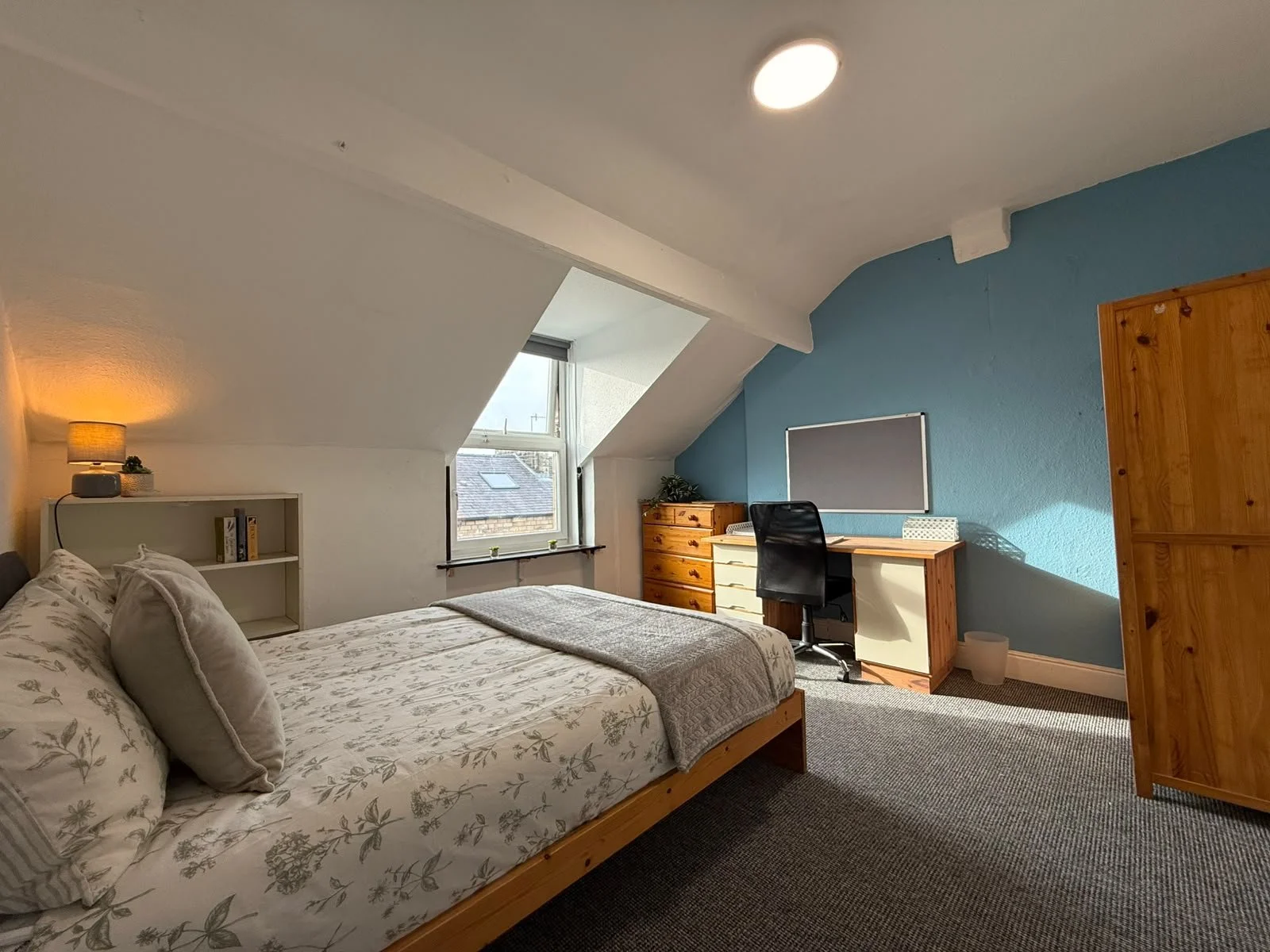 A cozy bedroom with a sloped ceiling, a bed with floral bedding, a nightstand with a lamp, a window with a view of rooftops, a wooden dresser, a desk with a black chair, a whiteboard, and an armoire.