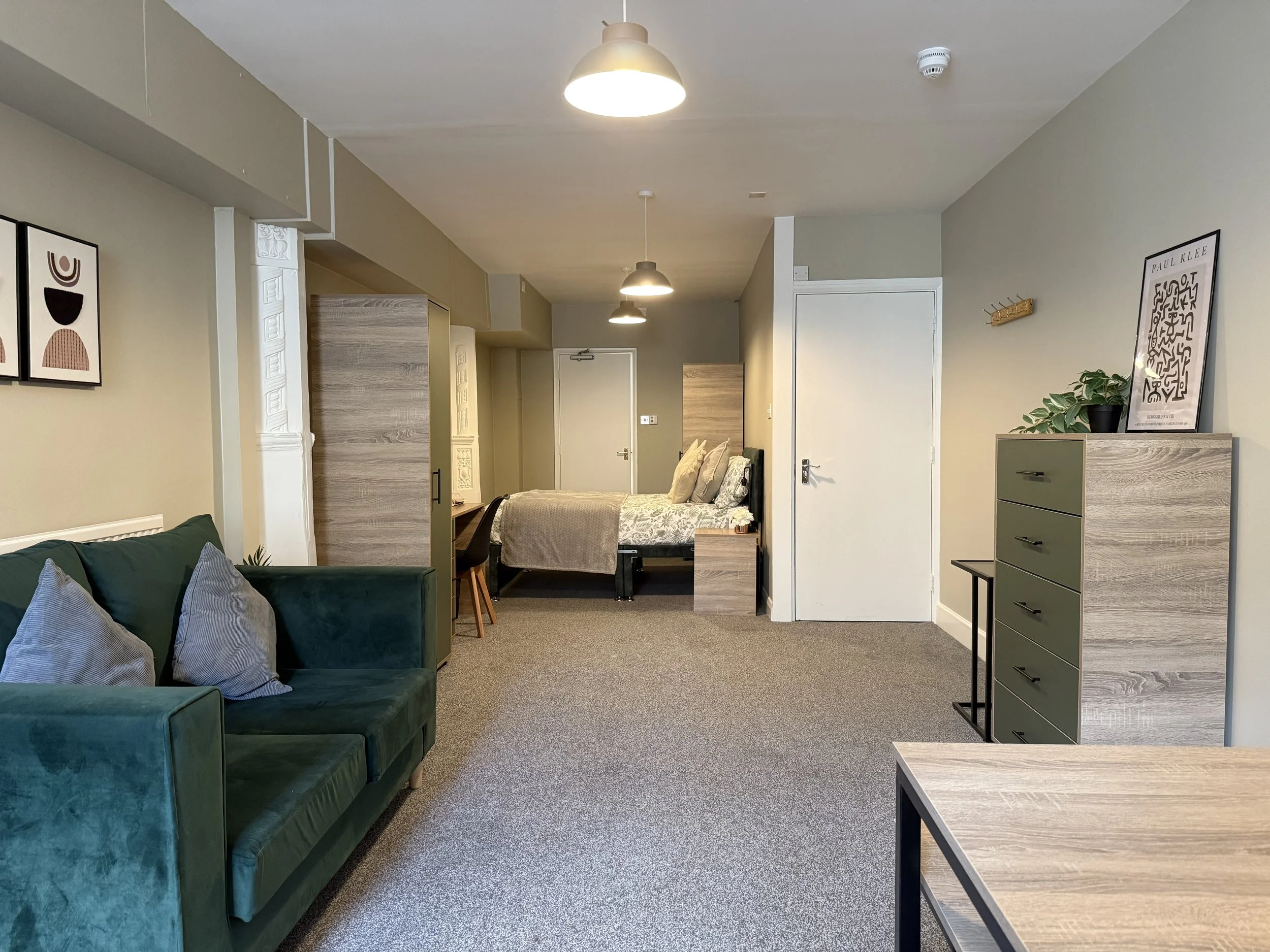 Interior of a modern studio apartment with a green couch, a bed with beige bedding, a dresser with artwork and a plant on top, and a small table. The room has beige walls, gray carpeting, and ceiling lights.