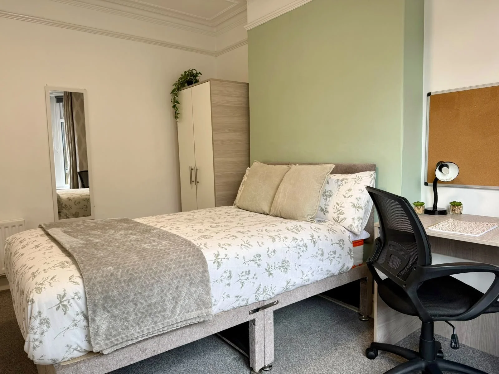 Bedroom with bed, pillows, green accent wall, wardrobe, desk with two small plants, corkboard, and office chair.
