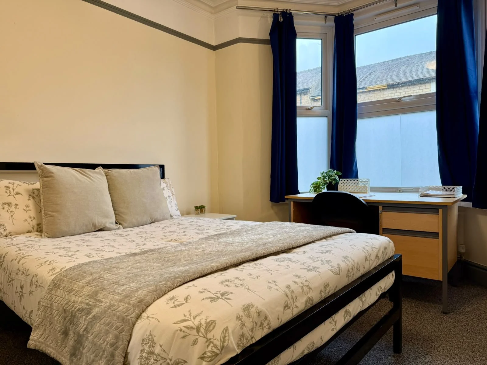 Clean bedroom with a bed, pillows, a desk, a chair, and windows with blue curtains, overlooking neighboring buildings.