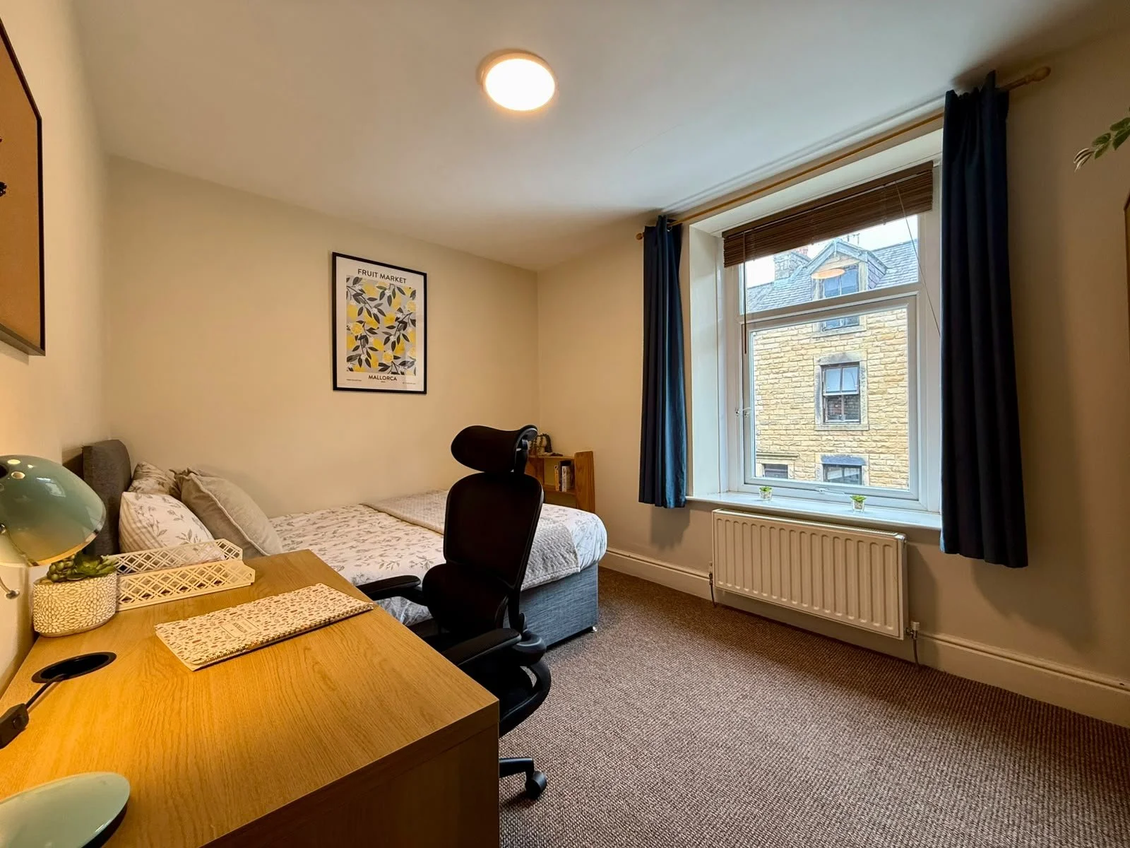A cozy bedroom with a bed, wooden desk, and office chair near a window with dark blue curtains. The room has a framed poster on the wall and a radiator under the window.