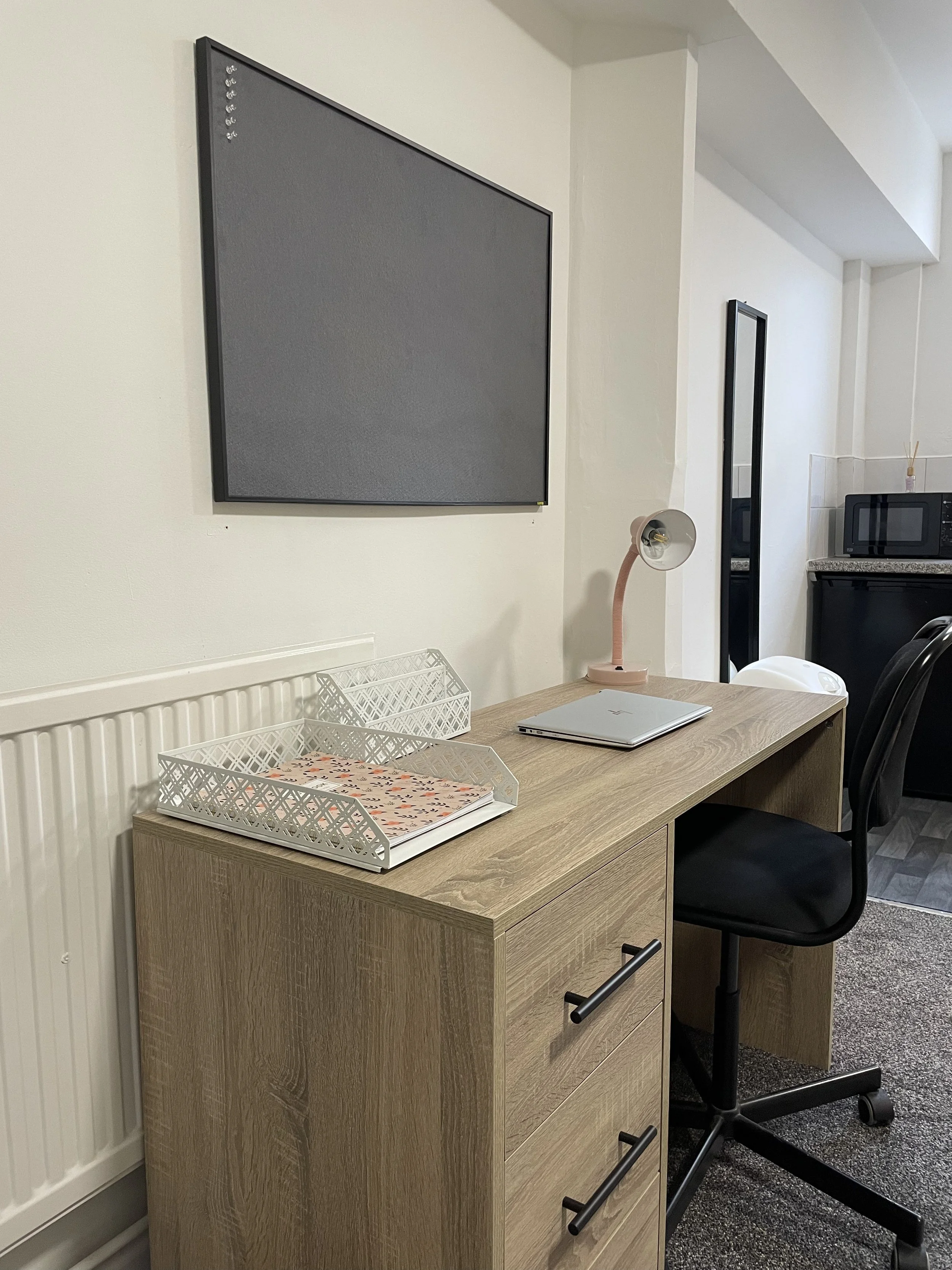 A wooden desk with three drawers, a pink desk lamp, a closed laptop, and a white decorative tray with paper inside. Behind the desk is a black office chair, a radiator, and a wall-mounted blackboard or corkboard. In the background, part of a kitchen