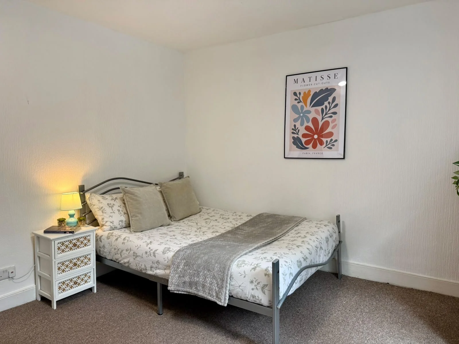 A bedroom with a bed, bedside table with a lamp and books, and a framed flower art print on the wall.