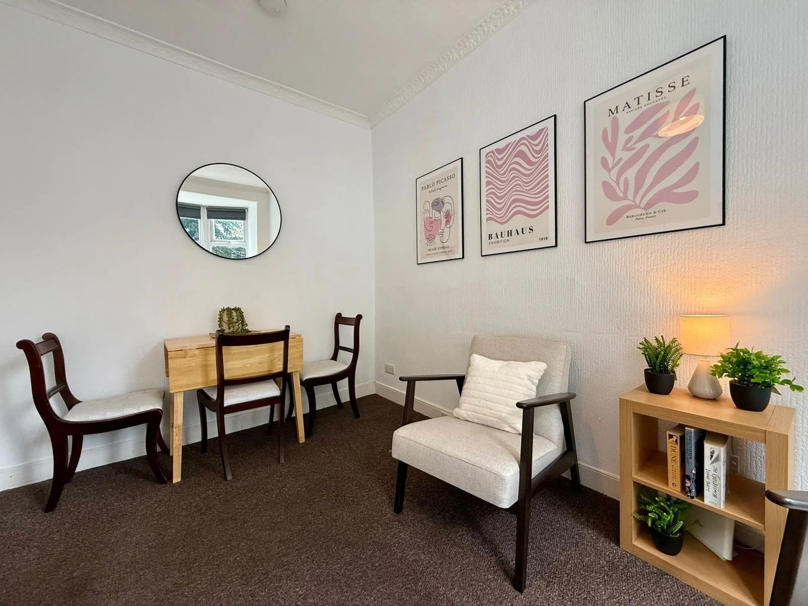 A cozy living room corner with a white armchair, a small wooden bookshelf with potted plants and books, a small side table with a lamp and more plants, three framed art prints on the wall, a mirror above a small wooden desk with two chairs, and a win