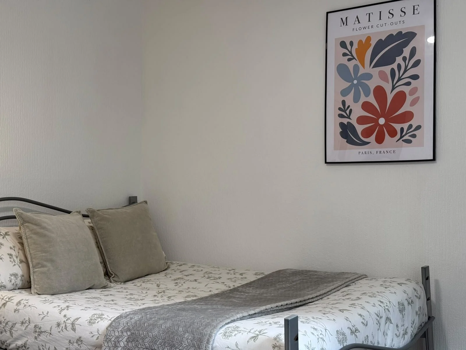 A neatly made bed with beige pillows and a floral patterned bedspread in a minimalistic bedroom. A framed art print titled 'Matisse Flower Cut-Outs' hangs on the white wall above the bed.