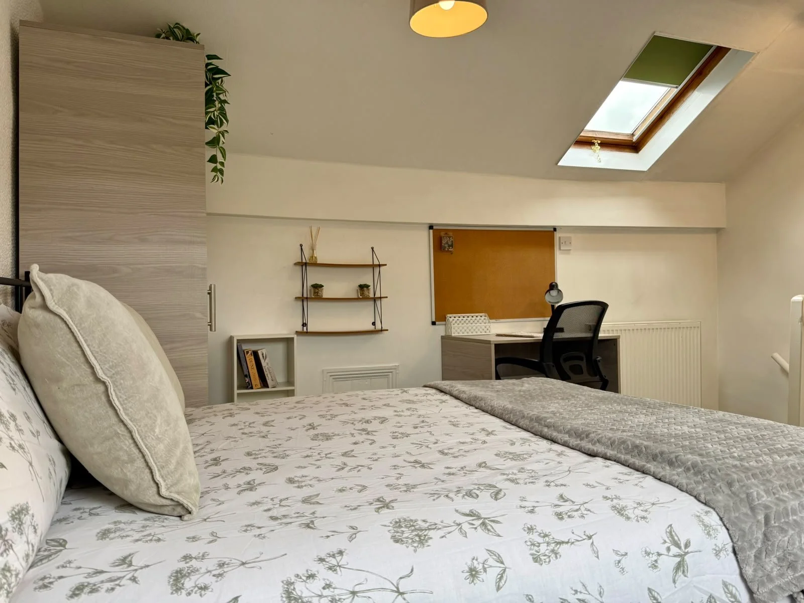 A cozy bedroom with a bed featuring floral bedding and plush pillows, a desk and office chair near a window, and decorative shelves on the wall beneath a skylight.