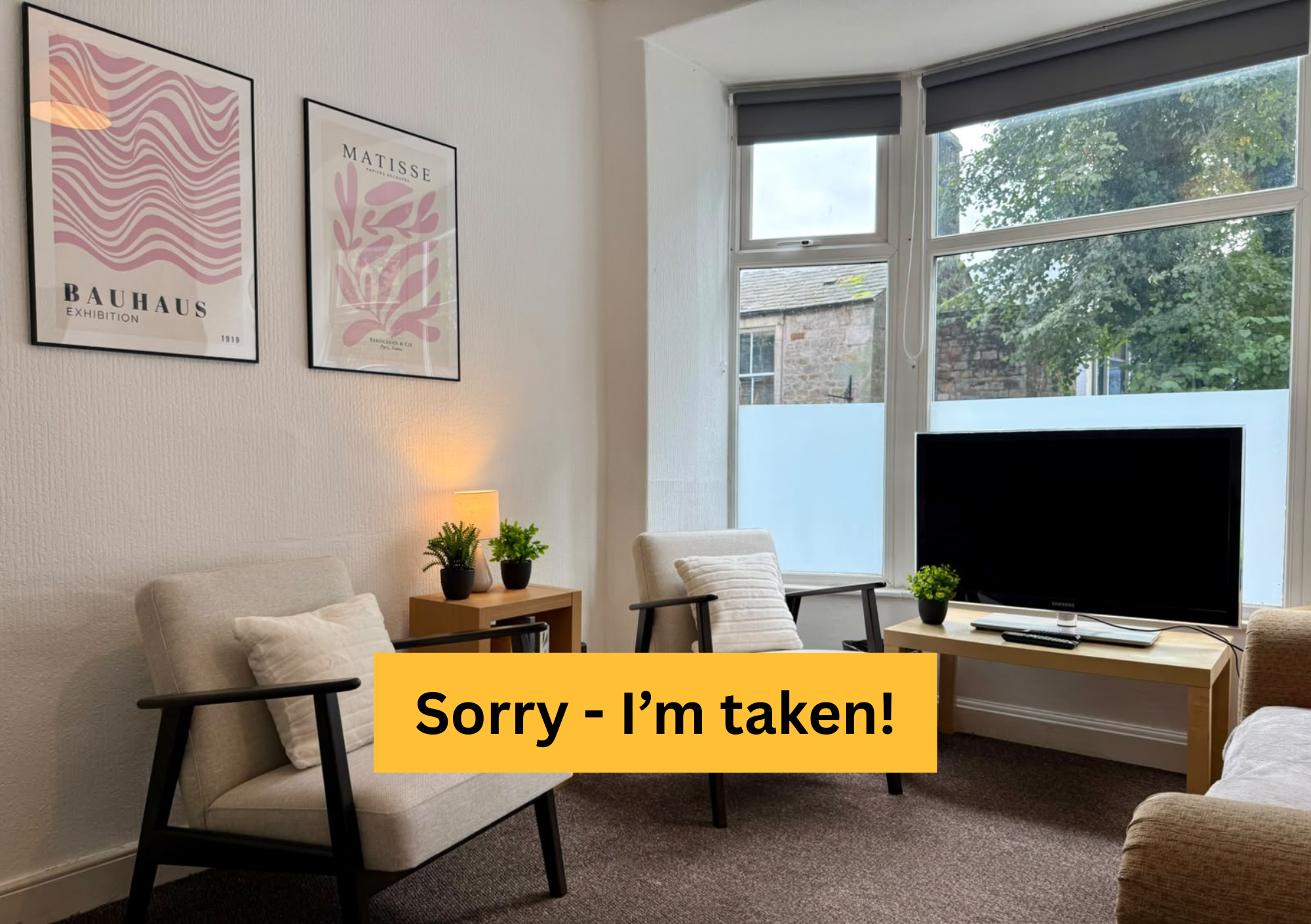 Living room with beige sofa and armchair, TV on a small table, window with trees outside, and framed posters on the wall.