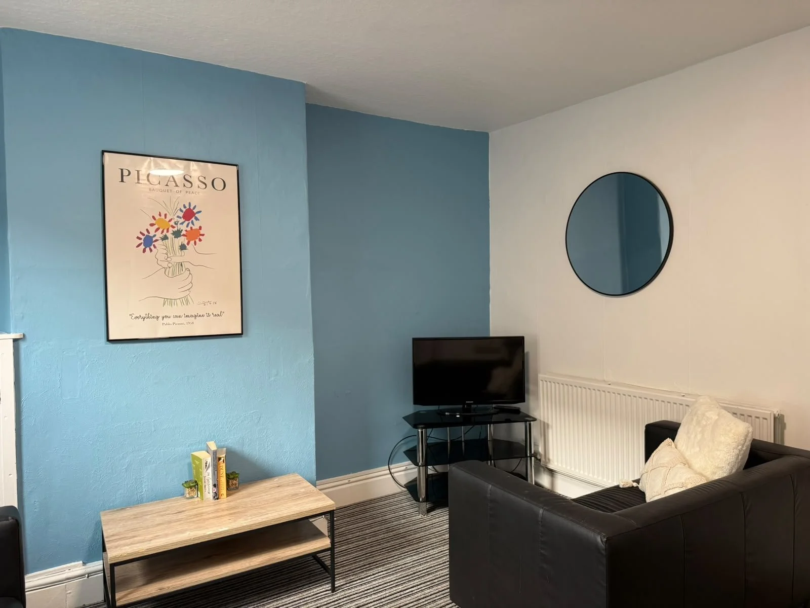 Living room with blue and white walls, a black sofa with cream cushions, a small TV on a black stand, a round mirror on the white wall, a framed Picasso poster, a light wood table with books and small plants, and a white radiator.