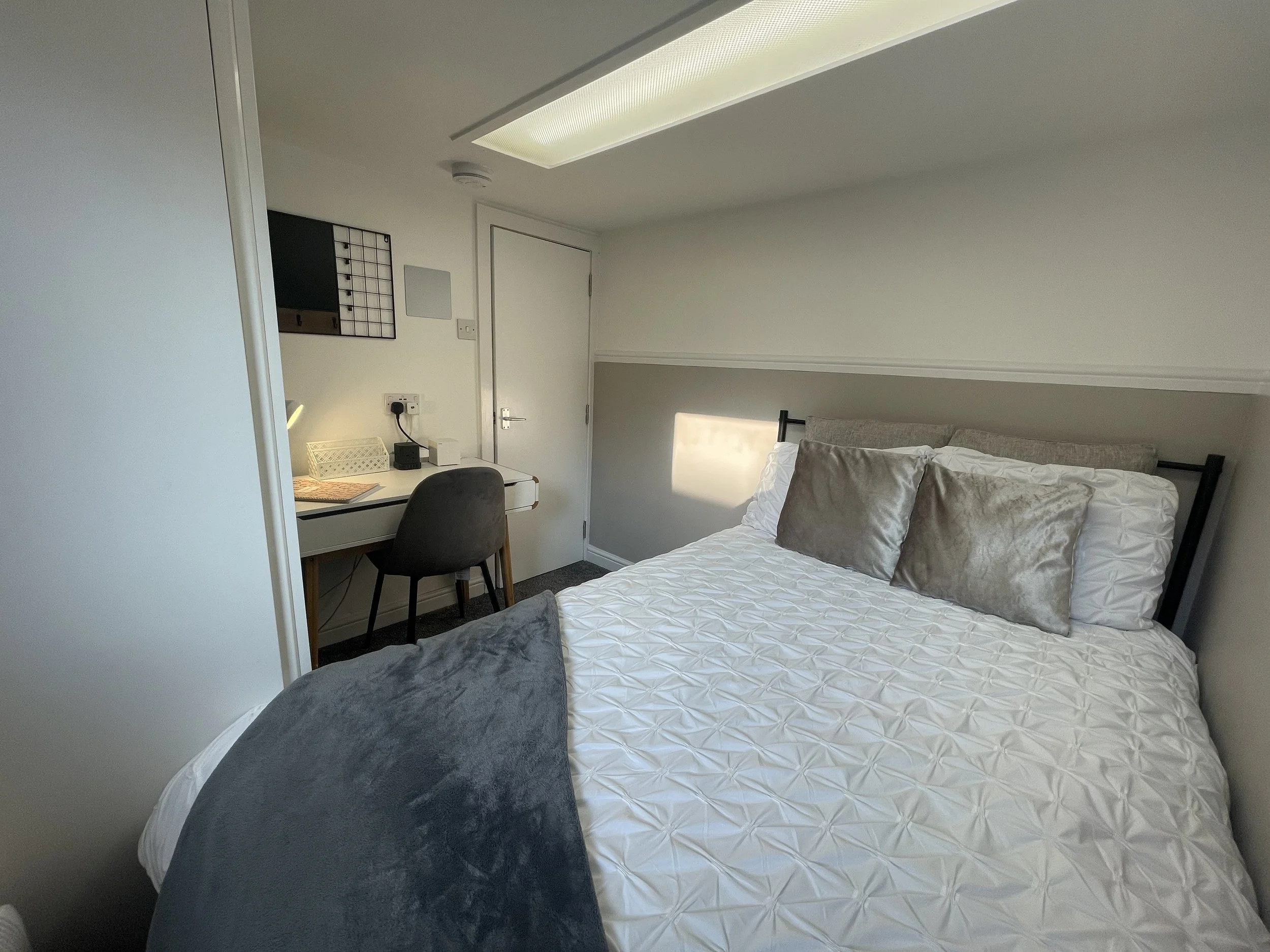 A bedroom with a bed dressed in white and gray pillows, a dark gray blanket at the foot of the bed, and a small workspace with a white desk, a gray chair, and a wall-mounted organizer. There is a door and a window allowing natural light.
