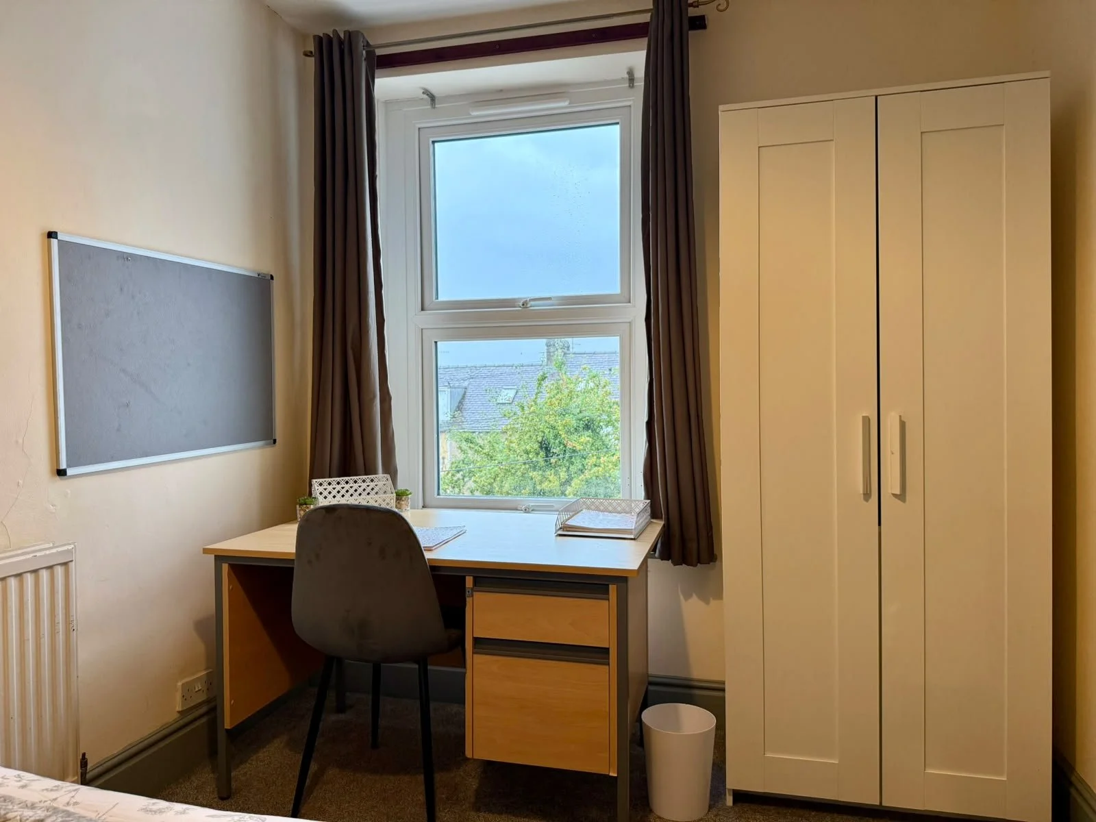 A small, tidy office with a desk and a black office chair. There is a window with dark curtains, a white cupboard on the right, and a blackboard on the left wall.