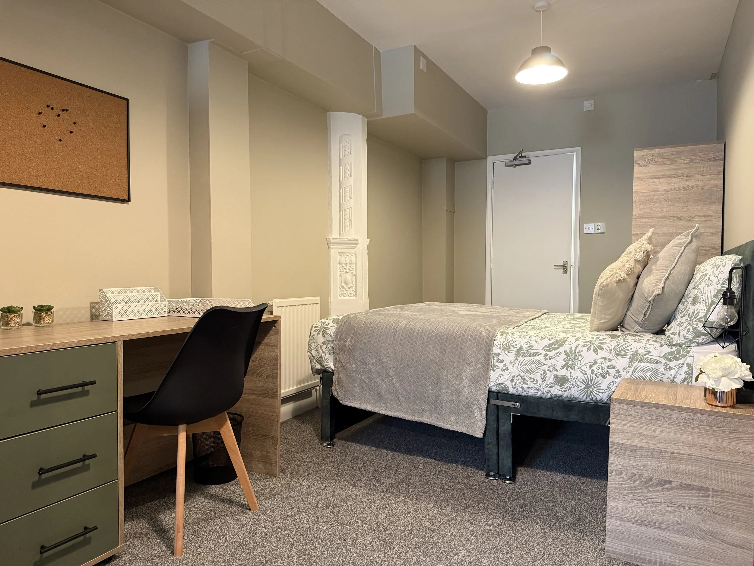 A modern, cozy bedroom with a bed featuring floral sheets and multiple pillows, a small bedside table with a flowers, a desk with decorations, and a black chair, with a radiator, a door, and minimalist decor in the background.