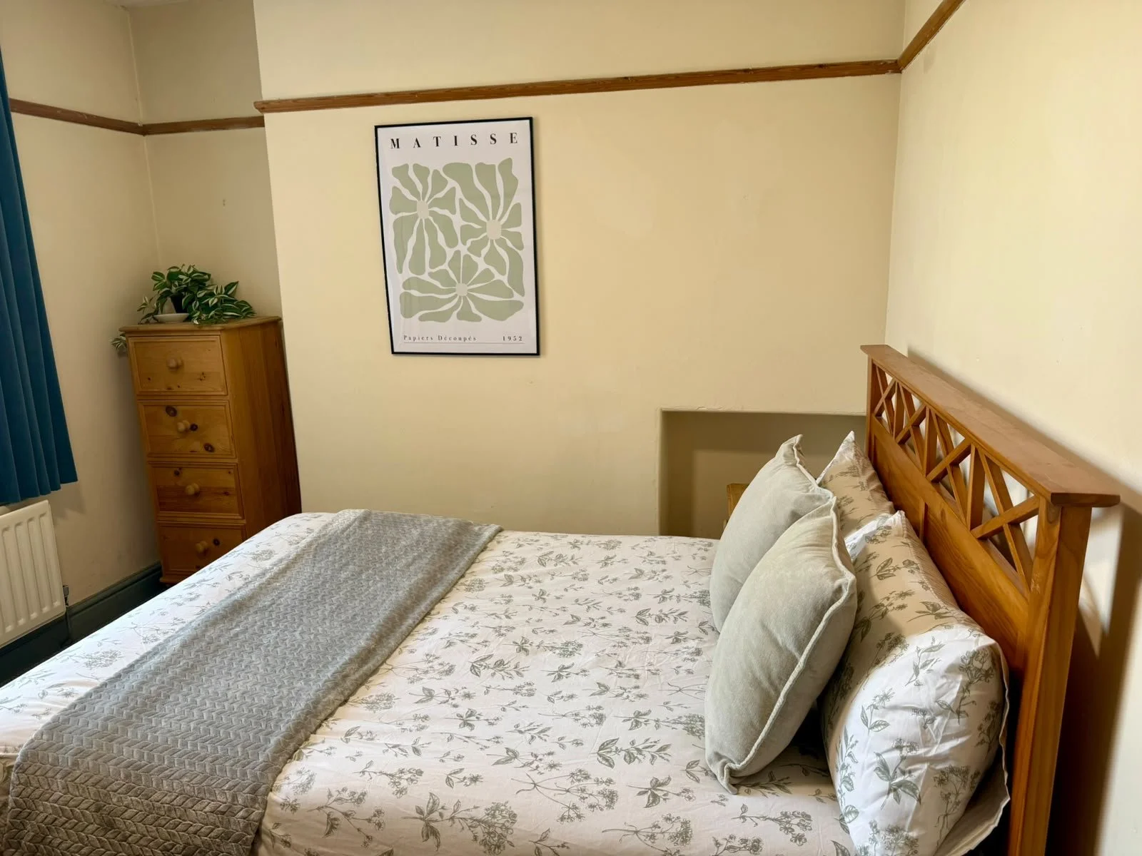 A bedroom with a wooden bed frame, floral bedding, and soft pillows. A wooden dresser with a houseplant in a white pot is placed in the corner, and a framed Matisse art print hangs on the cream-colored wall above the bed. A blue curtain partially cov