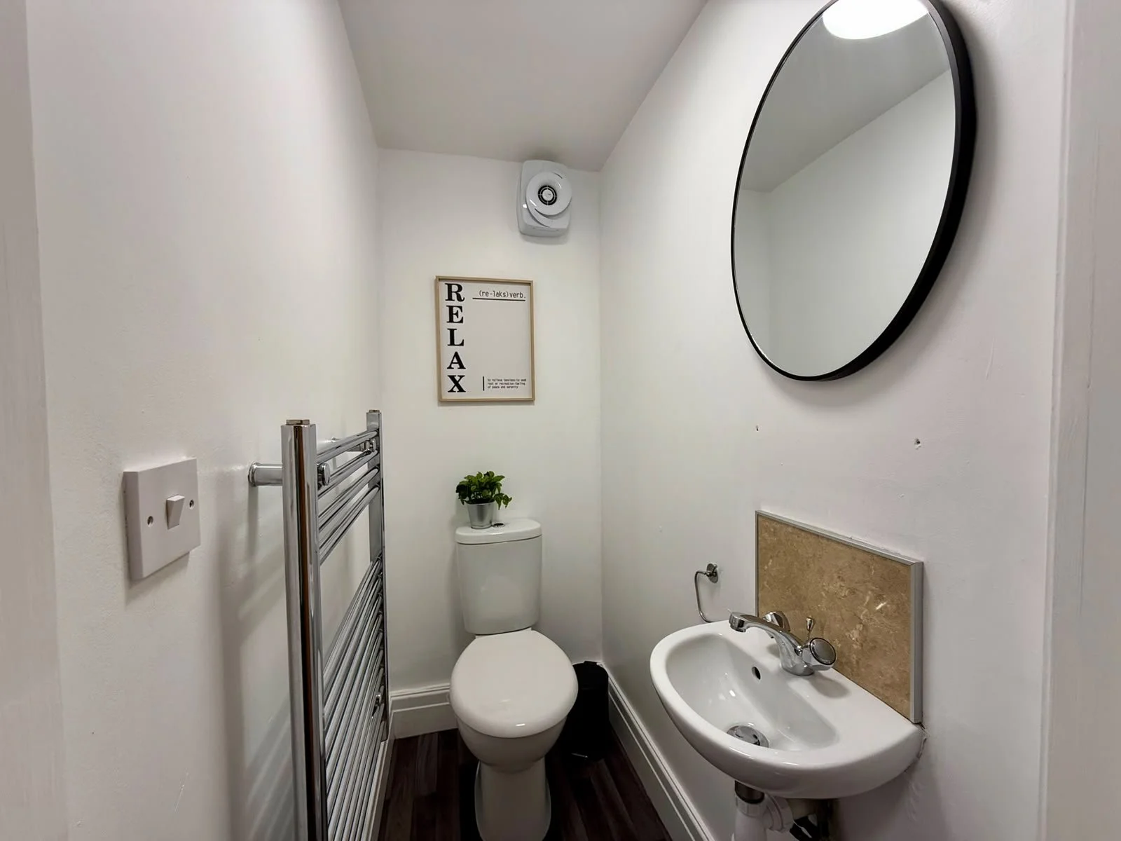 A small bathroom with a white toilet, small sink, round mirror, and a wall-mounted towel rack, decorated with a framed sign that says 'RELAX' and a small potted plant on the toilet tank.