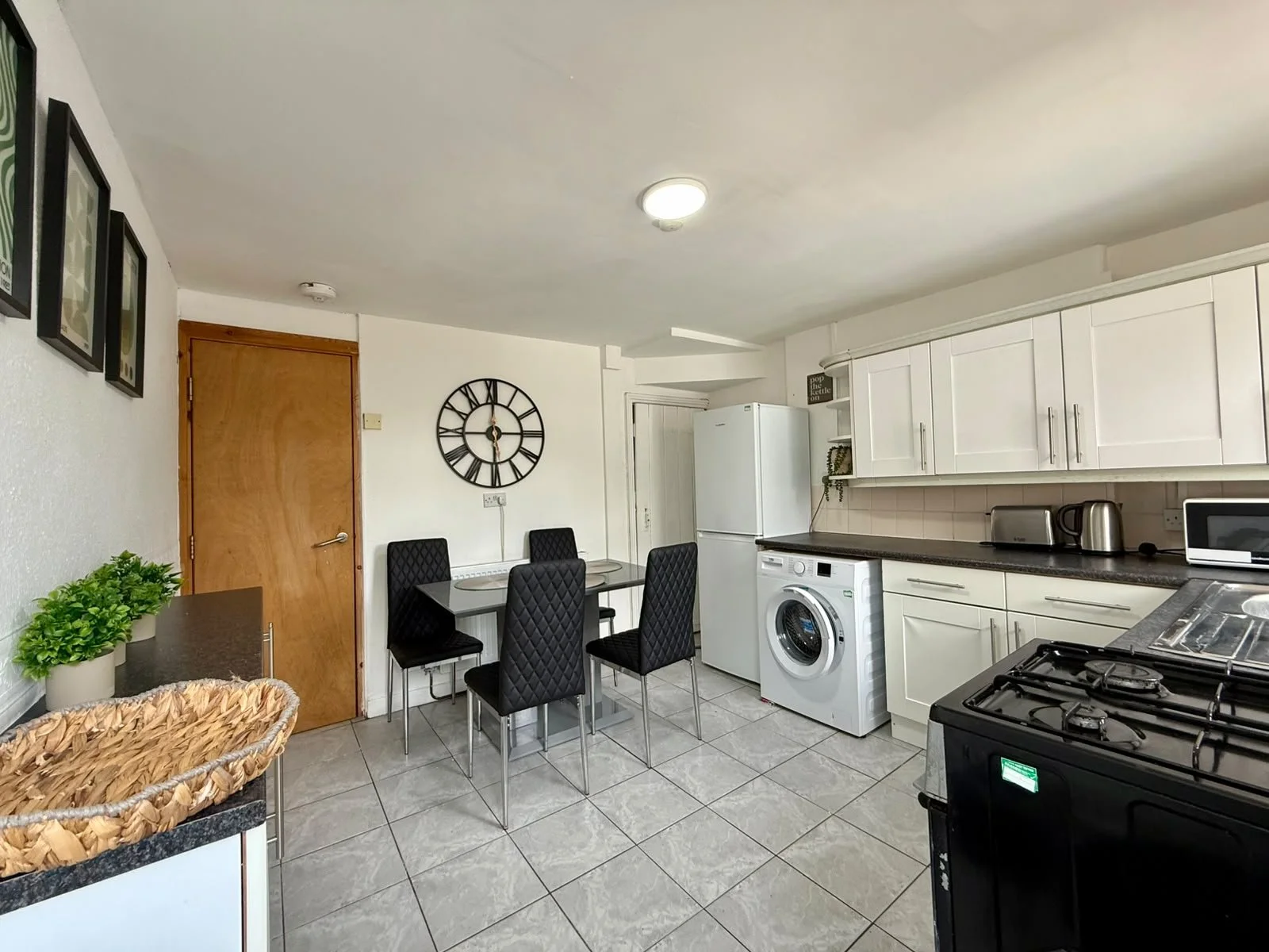 Kitchen and dining area with white cabinets, black countertops, a white refrigerator, washing machine, toaster, microwave, gas stove, and black chairs around a small dining table. There are wall decorations and potted plants.