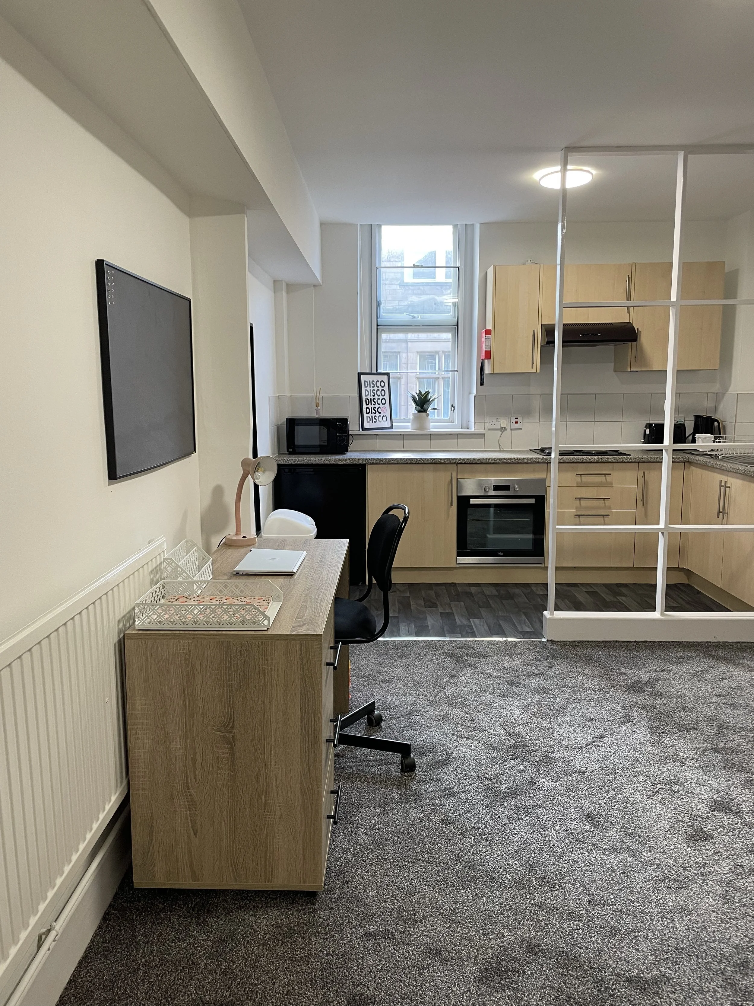 Small kitchen with wooden cabinets, black oven, microwave, toaster, and kettle. A desk with a laptop, lamp, and office chair. Window with a potted plant, framed art with the word "DISCO" repeated, and a glass partition.