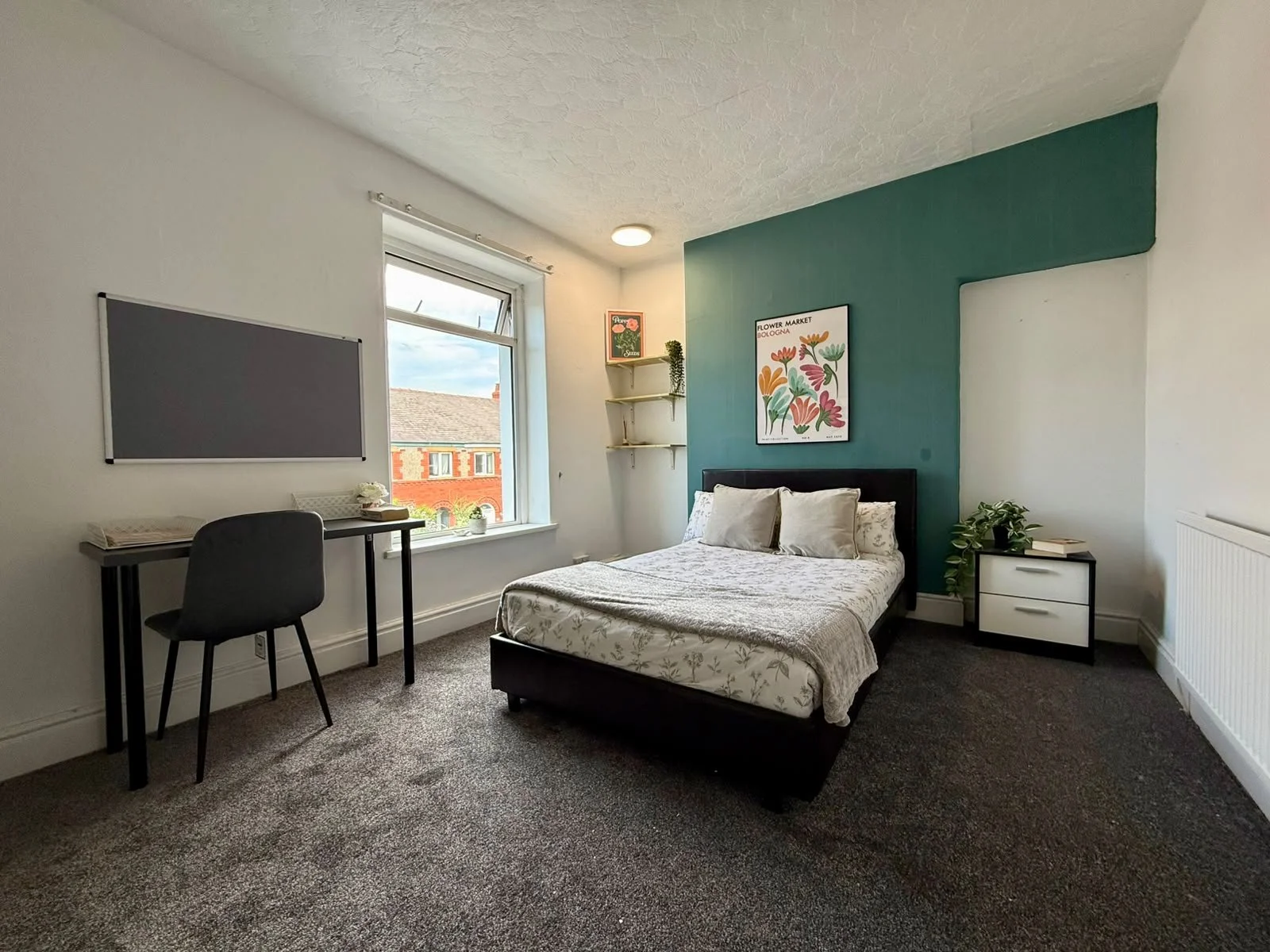 A bedroom with a bed against a teal accent wall, decorative pillows, and framed artwork. A window with a view of neighboring brick buildings nearby. A white nightstand with a green plant and books. A black desk with a chair and a gray chalkboard on t
