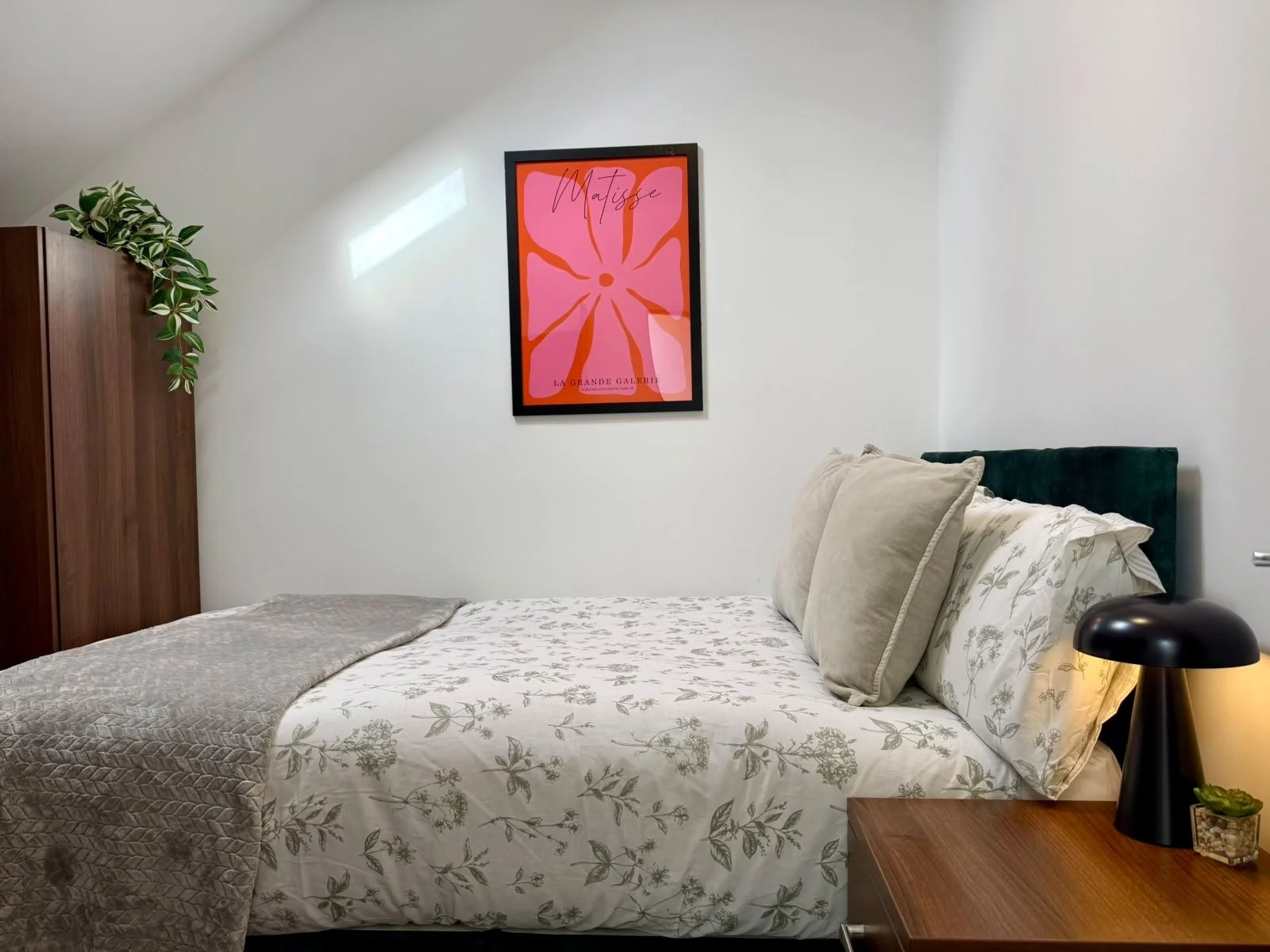 A cozy bedroom with a white floral bedspread, beige pillows, dark green headboard, wooden nightstand with a black lamp and a small plant, white walls, a framed pink and orange art piece, and a wooden wardrobe with a green leafy plant on top.