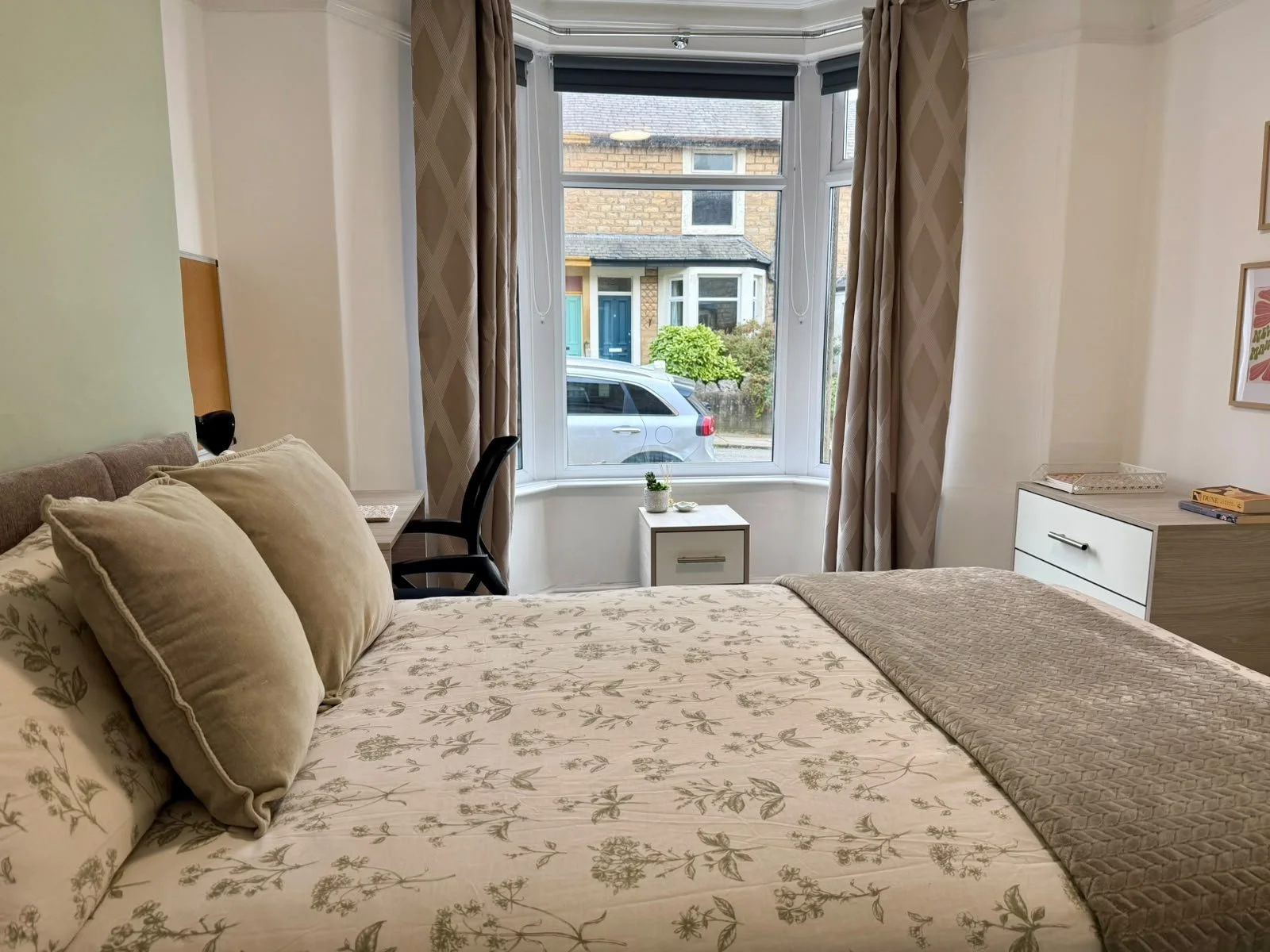 A cozy bedroom with a bed featuring floral-patterned bedding and beige pillows, a bedside table with a small potted plant, a black chair next to a desk, a white dresser with books, and a large bay window with beige curtains, overlooking a street with