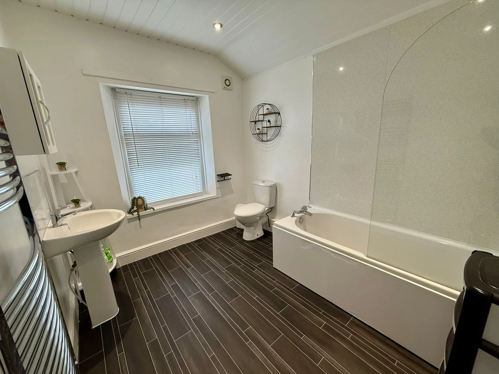 A modern bathroom with dark wood tile flooring, a white bathtub, a toilet, a pedestal sink, a window with blinds, a round wall shelf, and a heated towel rack.