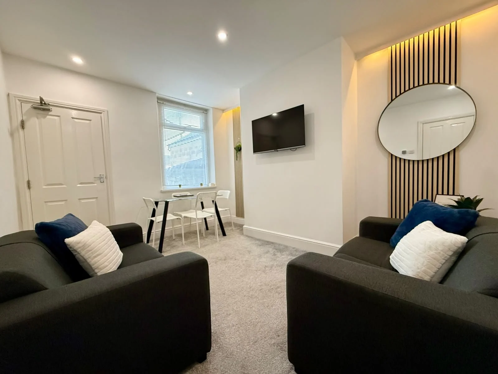 Modern living room with two black sofas with white and blue cushions, a wall-mounted TV, a round mirror on a decorative wooden panel, a small dining area with white chairs and a black table, and a window with blinds.
