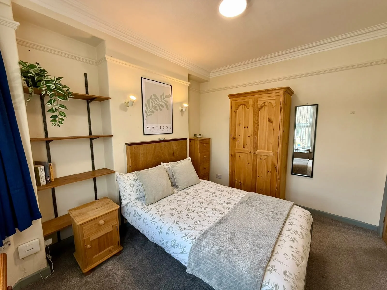 A cozy bedroom with a bed that has white and grey floral bedding, a wooden headboard, and pillows. To the left, there are wooden shelves with a plant and some books. On the right, a wooden wardrobe stands next to a mirror on the wall. The room has cr