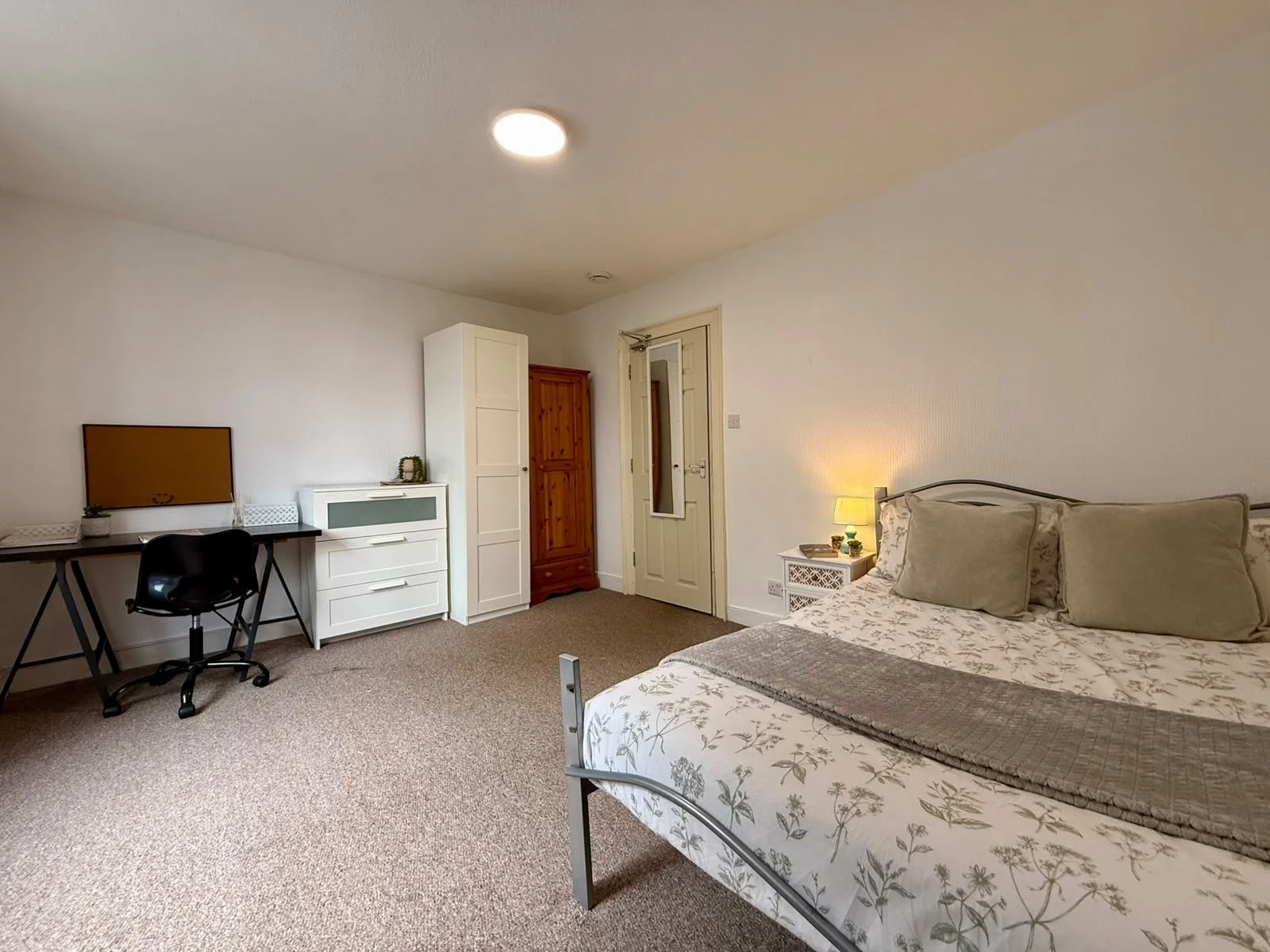 A cozy bedroom with a bed, nightstand, lamp, wardrobe, and a small workspace with a desk and chair, carpeted floor, and a plain white wall.