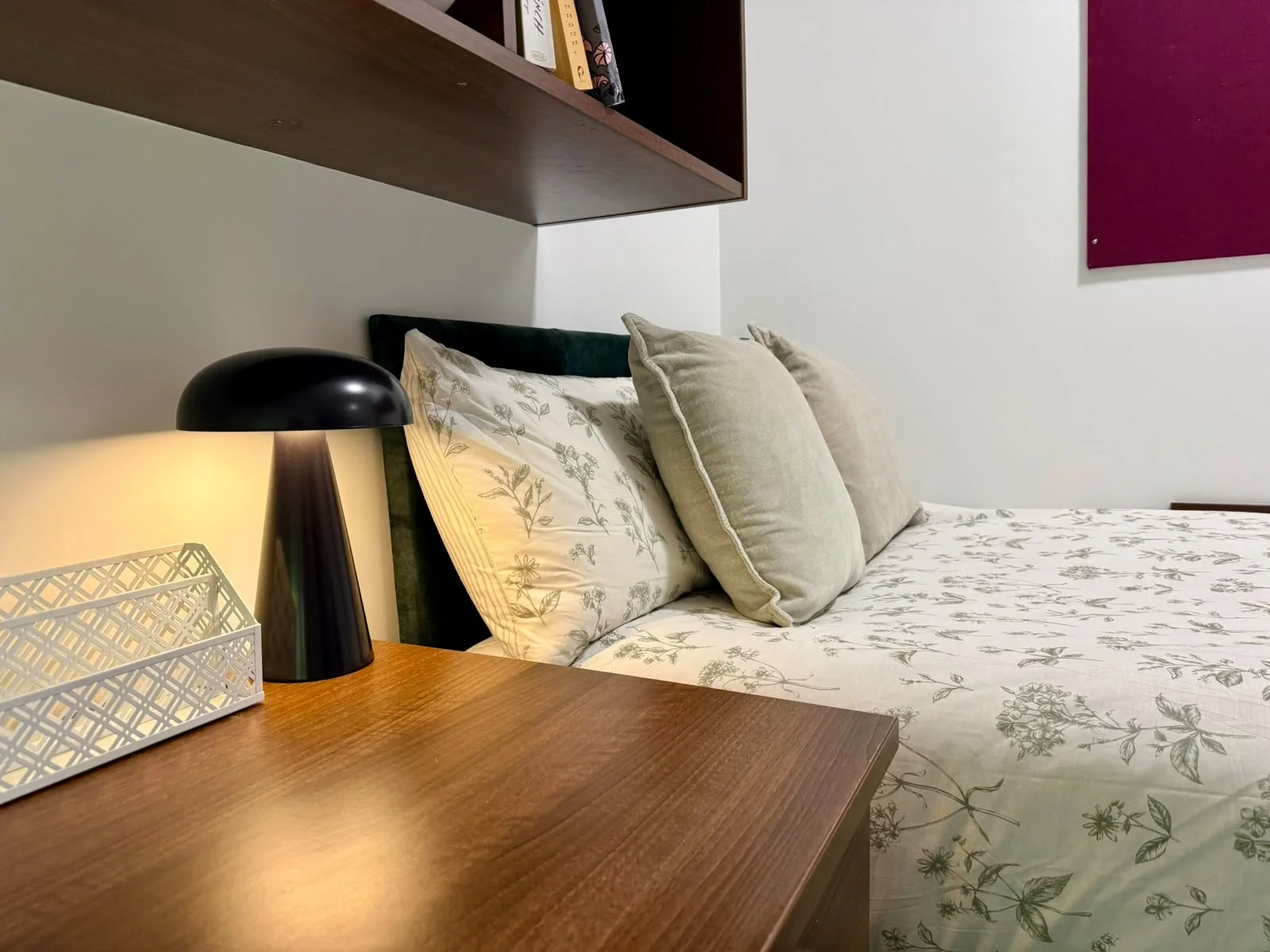 A neatly made bed with floral sheets and several beige pillows in a cozy bedroom. A wooden nightstand holds a black bedside lamp and a decorative white basket. A dark green headboard and a wall-mounted shelf with books are visible. Part of a purple w