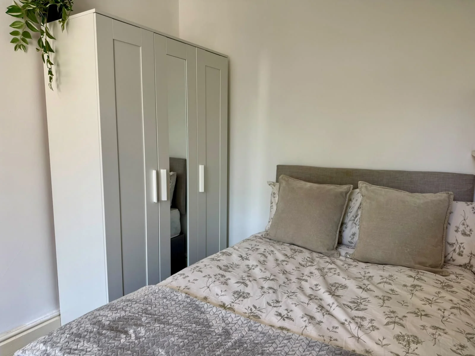 A bedroom with a bed covered in floral and gray quilted bedding, three beige pillows, a gray headboard, a white wall, and a gray wardrobe with a mirror door. A green plant is hanging from the ceiling on the left side.