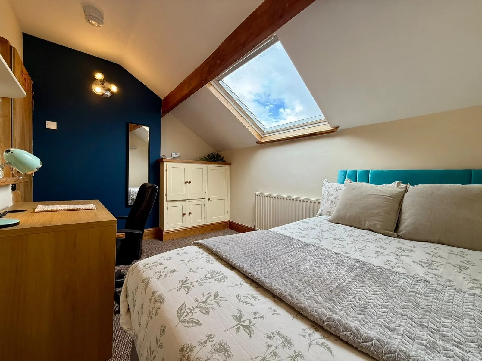 A bedroom with a skylight window showing a blue sky with clouds, a bed with floral bedding and pillows, a desk with a lamp, a black office chair, a white cabinet, and a mirror, with a wooden beam and beige walls.