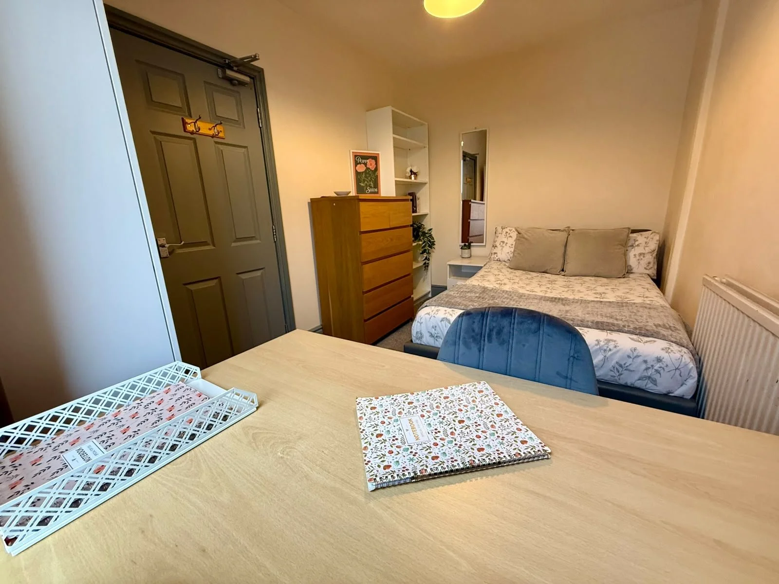 Cozy bedroom with bed, pillows, chest of drawers, mirror, and table with notebook and tray, next to white wardrobe and door.
