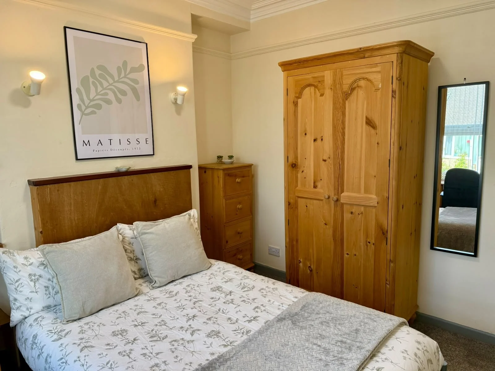 A cozy bedroom with a bed featuring light-colored floral patterned bedding and pillows, a wooden headboard, a small wooden bedside table, a large wooden wardrobe, a hanging mirror, wall-mounted lights, a framed poster of a leaf with the word 'Matisse