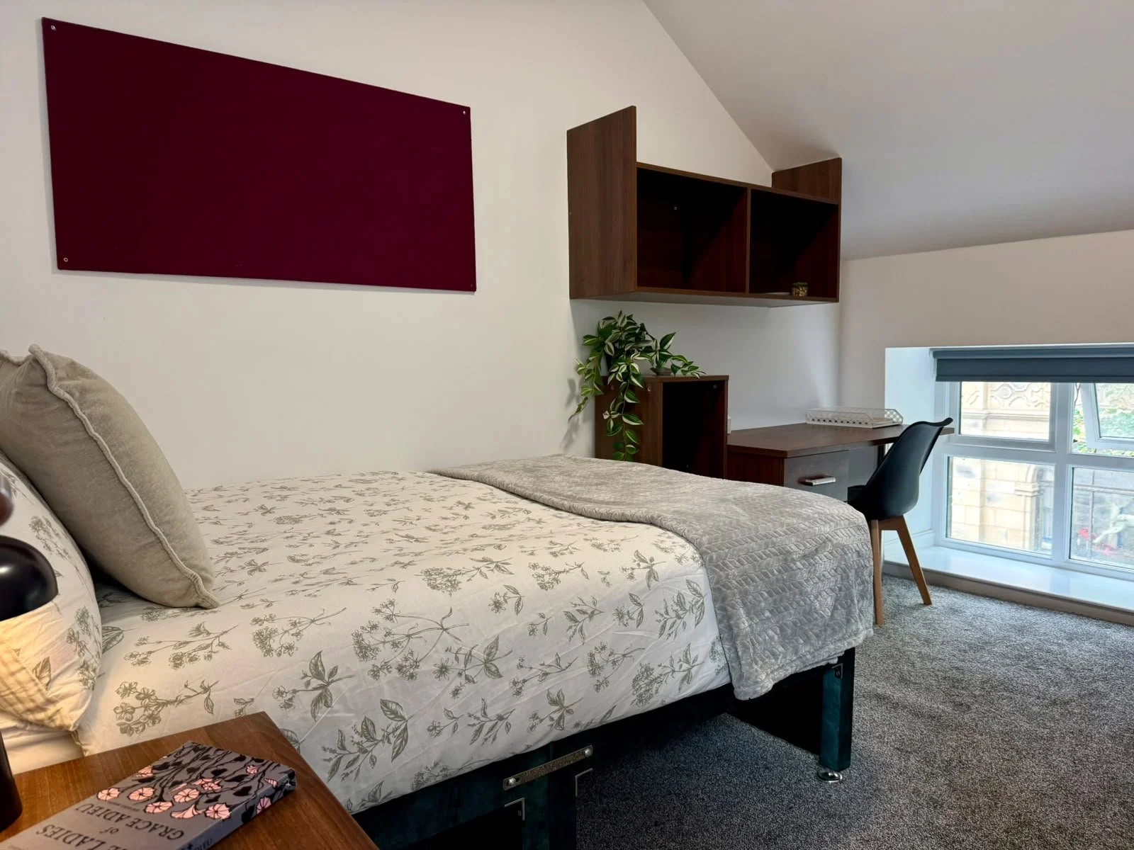 A cozy bedroom with a bed, a desk, a chair, and a window with a view of building and trees outside.
