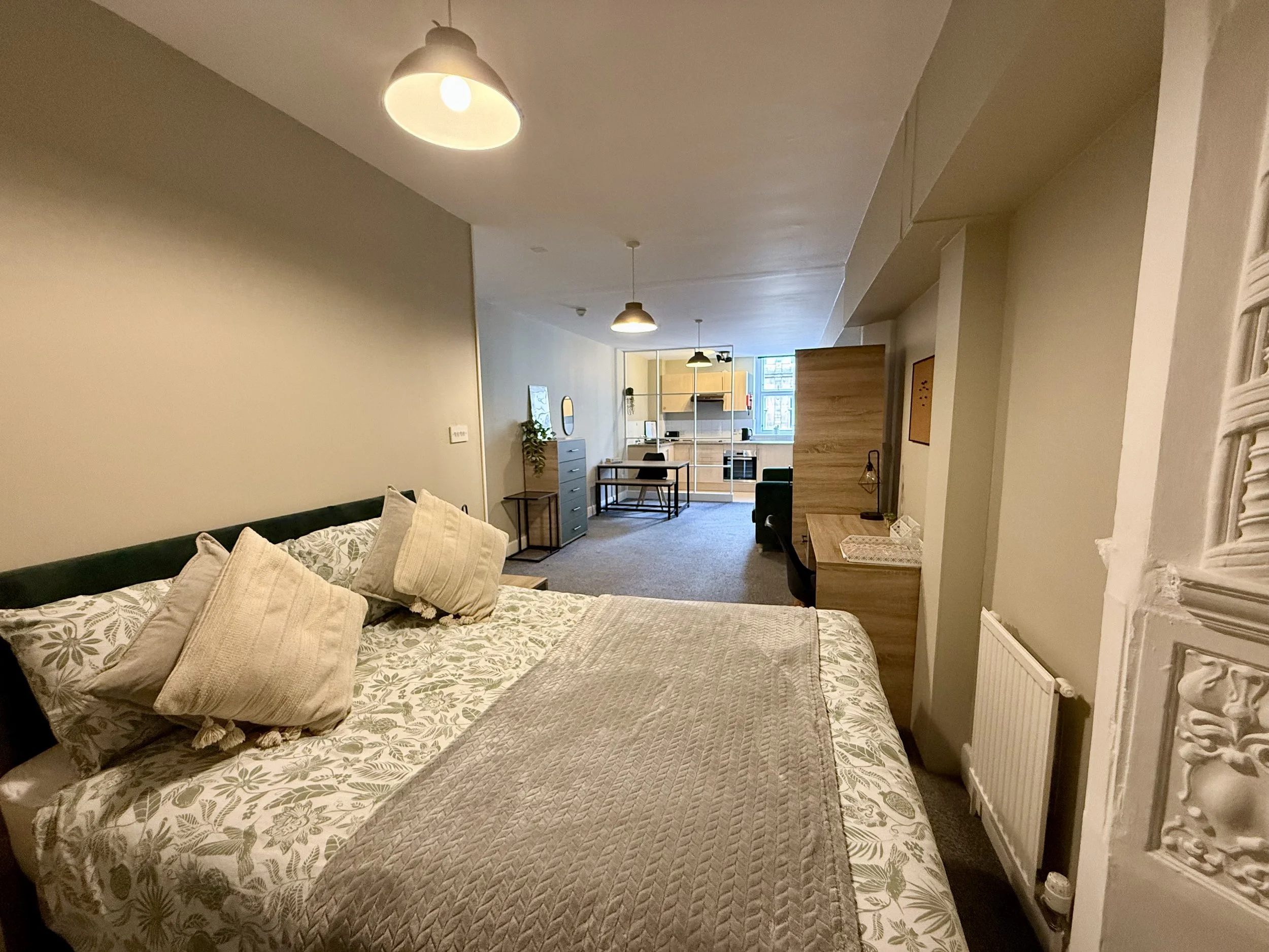 A bedroom with a bed covered in floral-patterned bedding and several pillows, a radiator on the wall, and a view into a living area with furniture and a kitchen in the background.