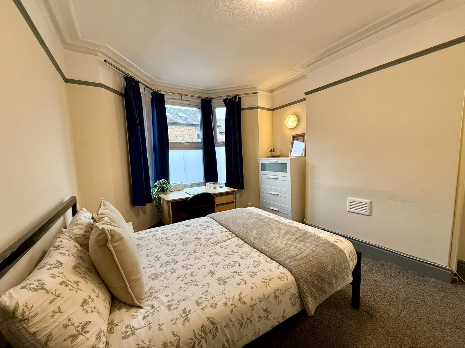 A cozy bedroom with a bed, pillows, a window with dark curtains, a desk with a chair, a white dresser, a small plant, and decorative items. The room has beige walls with gray trim and a carpeted floor.