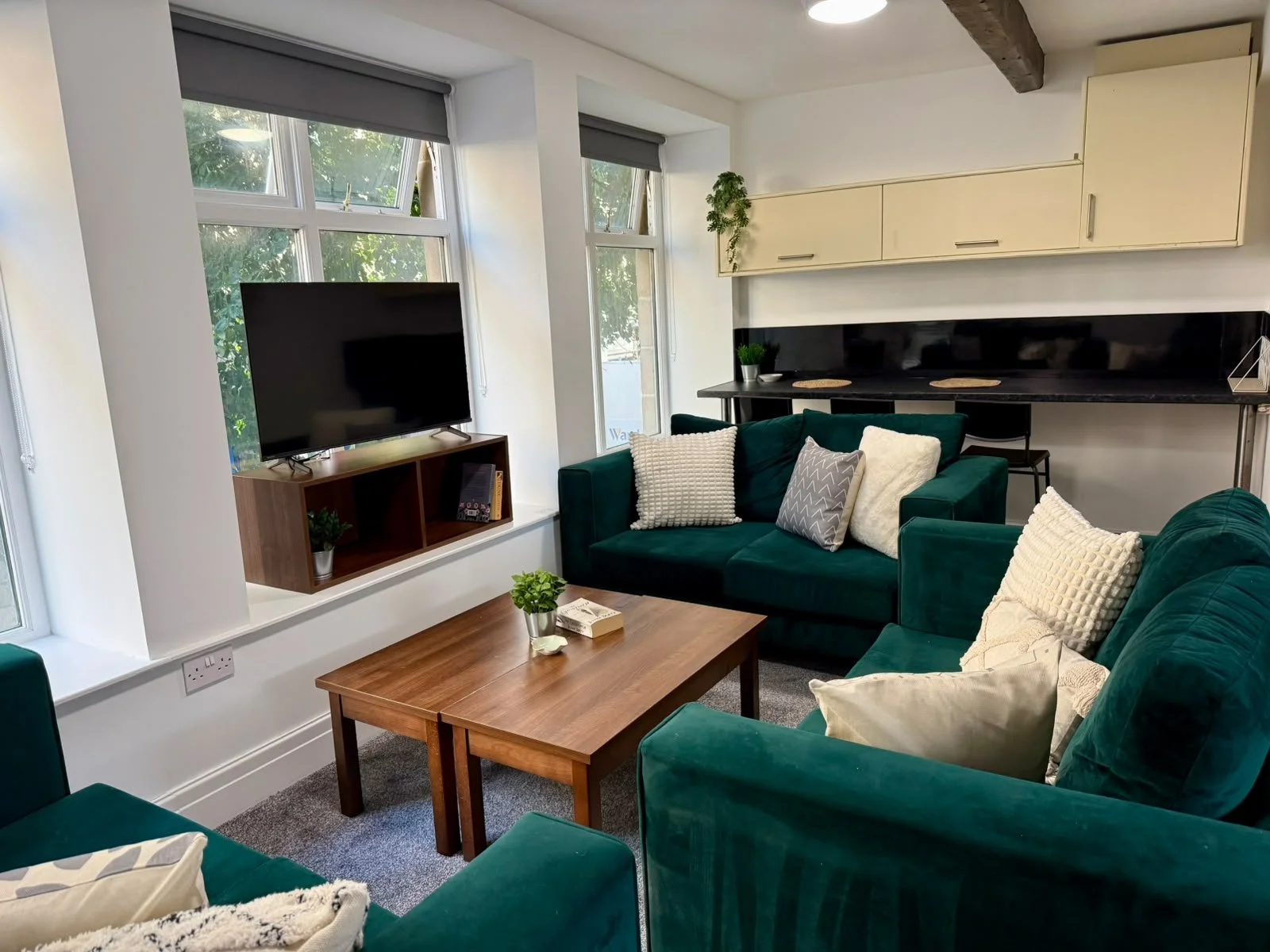 Living room with green sofas, a wooden coffee table, a TV on a wooden stand, large windows with gray blinds, and decorative pillows. There are houseplants and a black countertop with placemats and a barstool in the background.