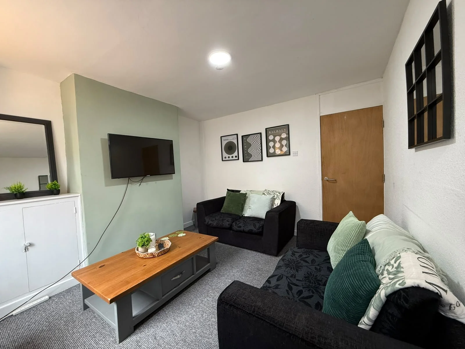 Living room with a black sofa, green and white pillows, a wooden coffee table with small plants, a wall-mounted TV, and framed artwork, with a wooden door and wall decor.