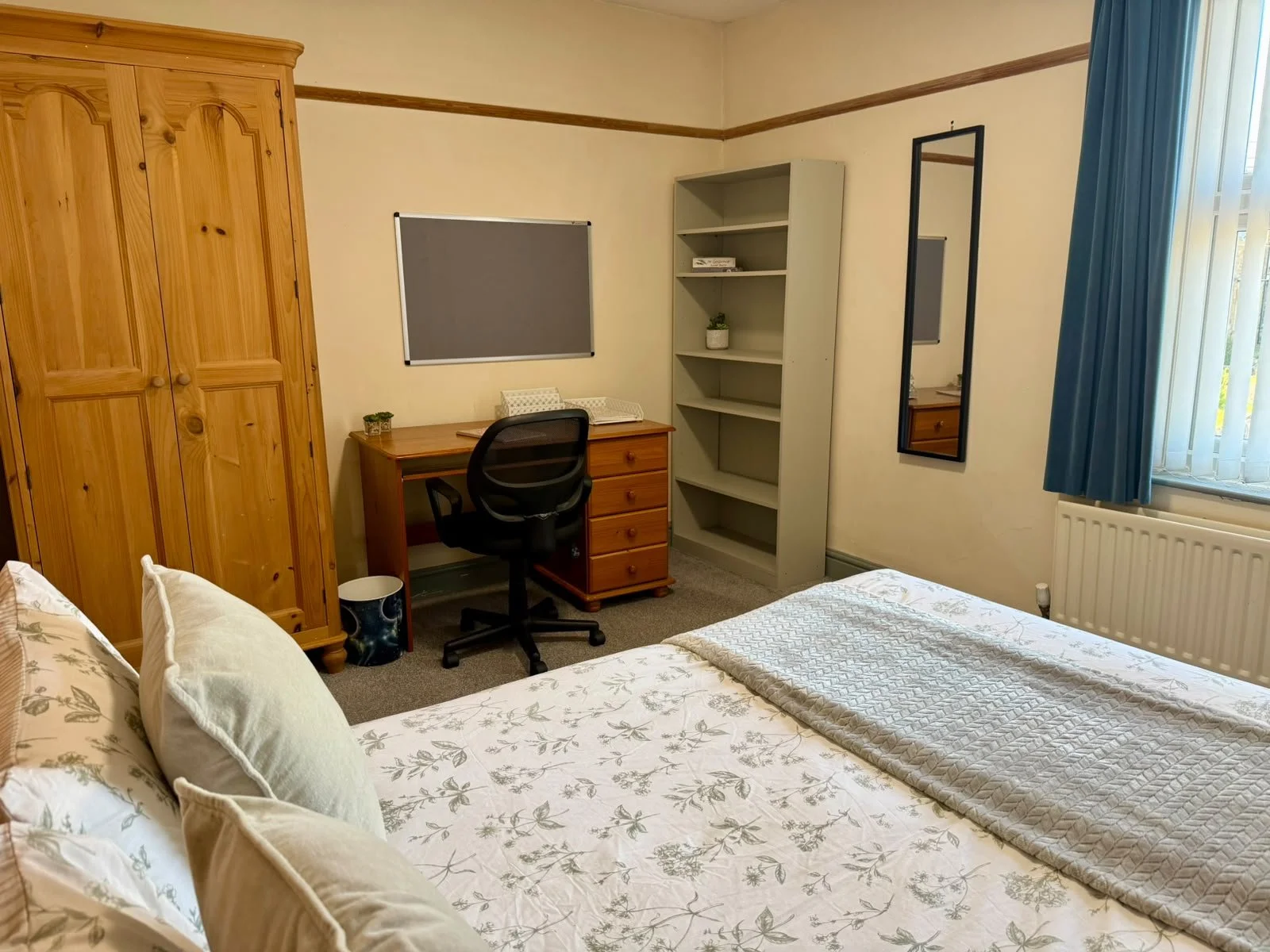 A cozy bedroom with a wooden wardrobe, a bed with floral-patterned linens, a window with blue curtains, a mirror on the wall, a small desk with a chair, a bookshelf, and some small decorative items.