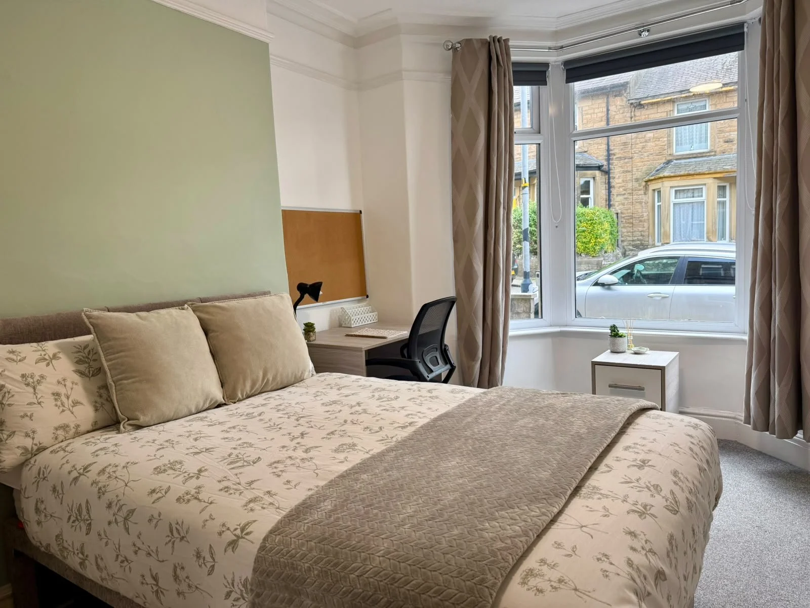 A cozy bedroom with a bed, beige floral bedding, pillows, a window with curtains, a small white nightstand with a plant, and a desk with a chair near the window.