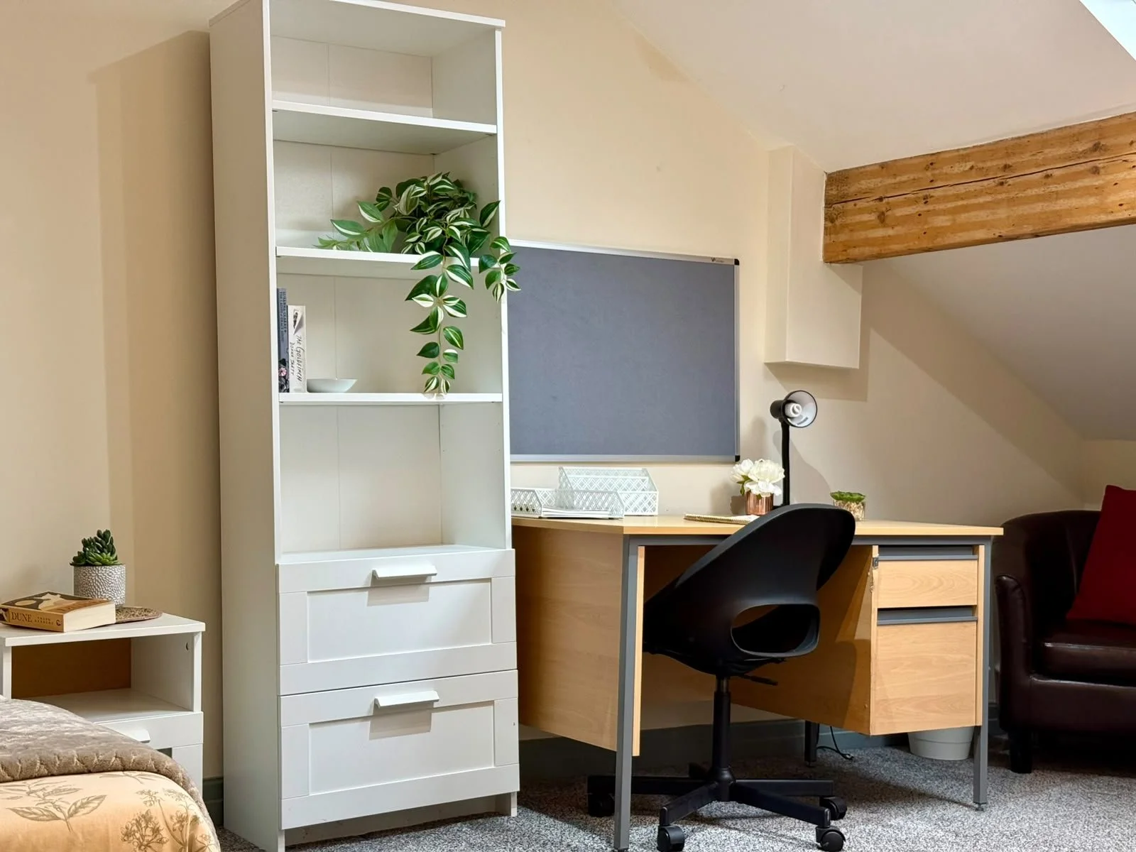 Home office in an attic with beige walls. A white bookshelf with a green plant, some books, and decorative objects. A wooden desk with a laptop, a flower vase, and a desk lamp. A black office chair and a small white table with a plant and a book. A g