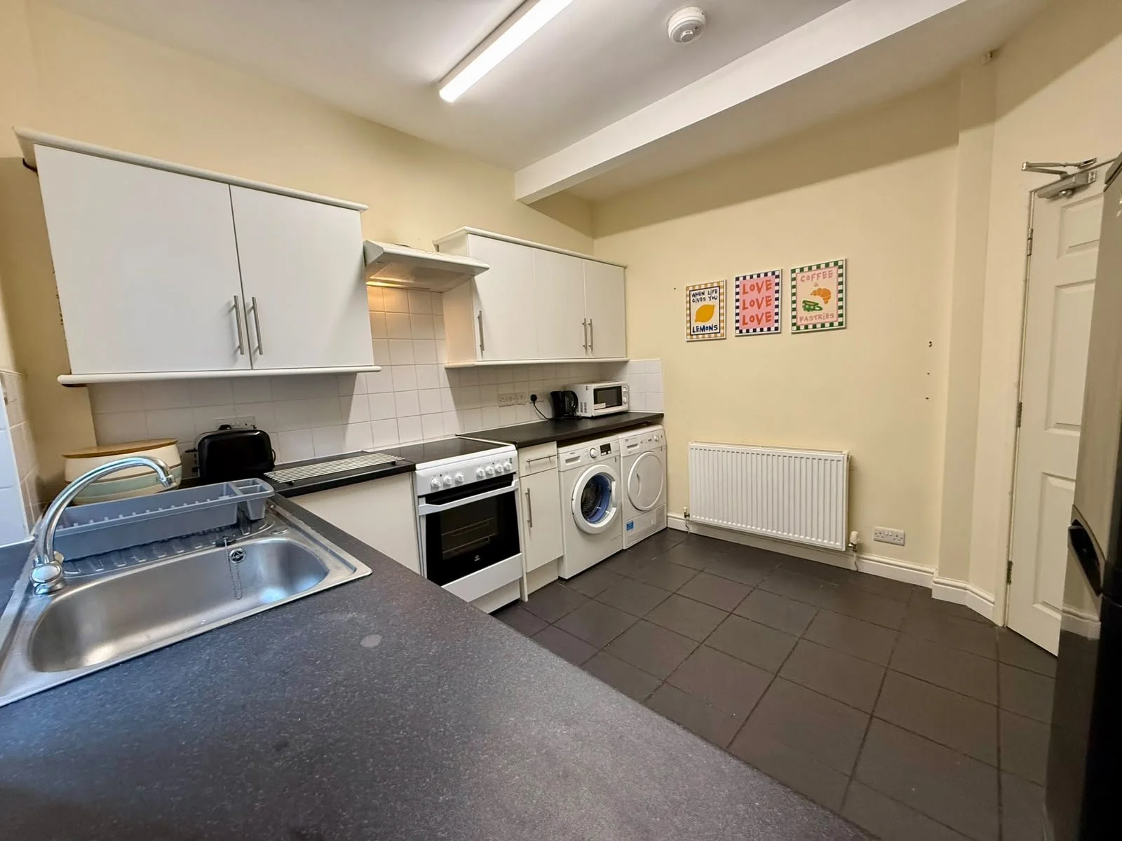 A kitchen with white cabinets, a black countertop, a stainless steel sink, a white oven, washing machine, and microwave. Decorated with three colorful framed art pieces on the wall.