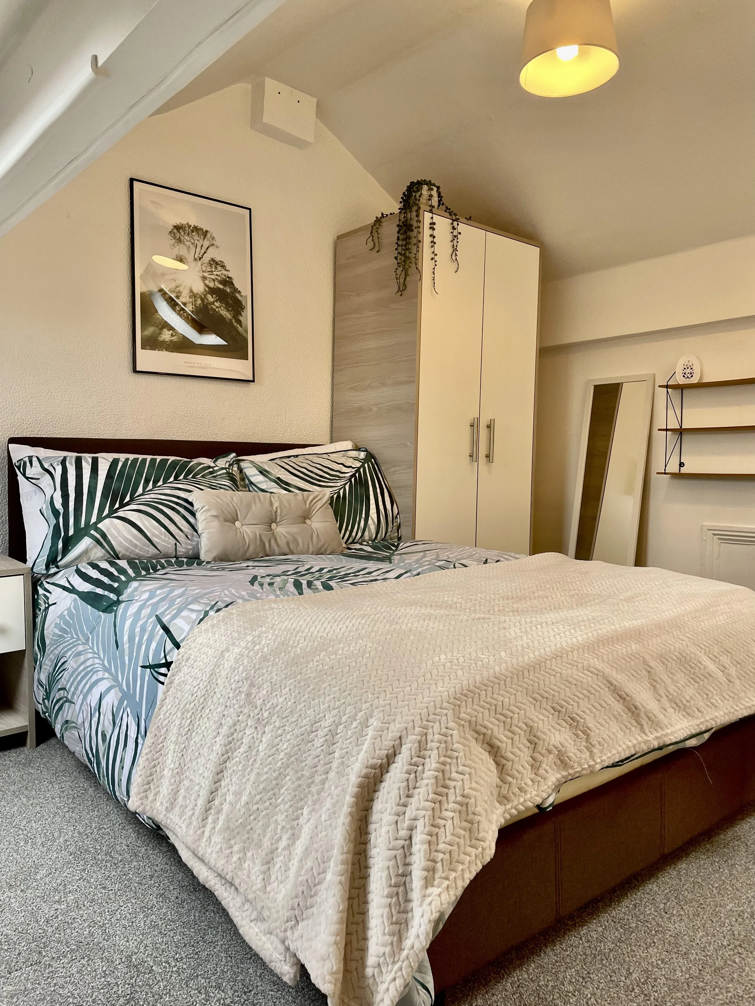 A bedroom with a bed featuring palm leaf patterned bedding, a beige pillow, a nearby white nightstand, a framed tree poster on the wall, a wardrobe with a plant on top, a mirror leaning against the wall, and a wall-mounted shelf.