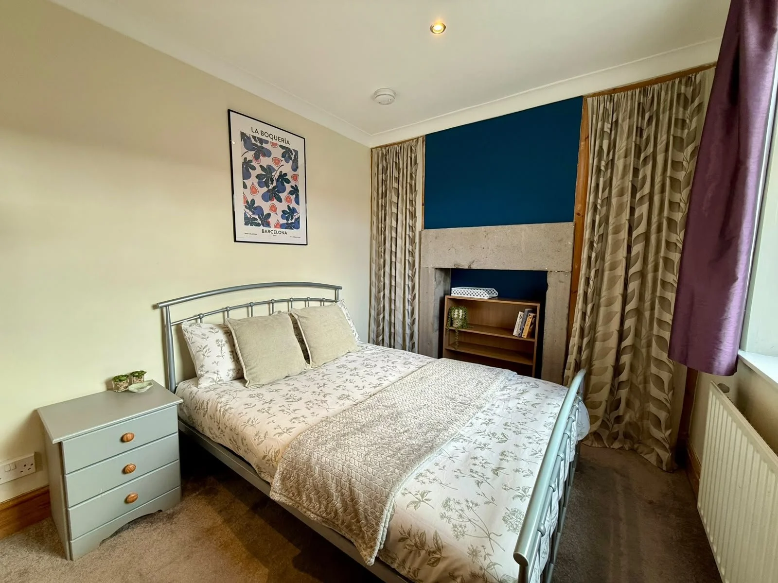 A bedroom with a bed, nightstand, and a fireplace with a blue accent wall. Curtains and a picture on the wall are also visible.