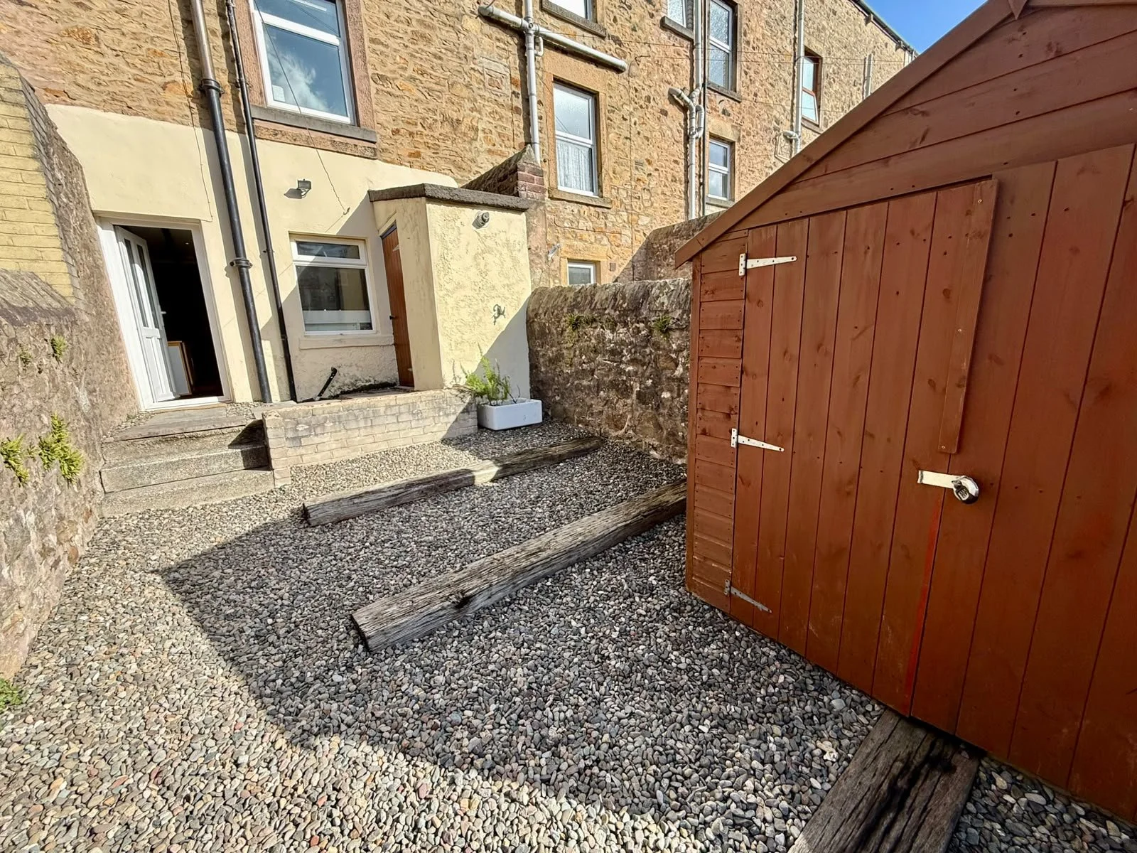 Backyard with gravel ground, stone steps leading to a door, a red wooden shed with a padlock, and a stone wall with a house in the background.