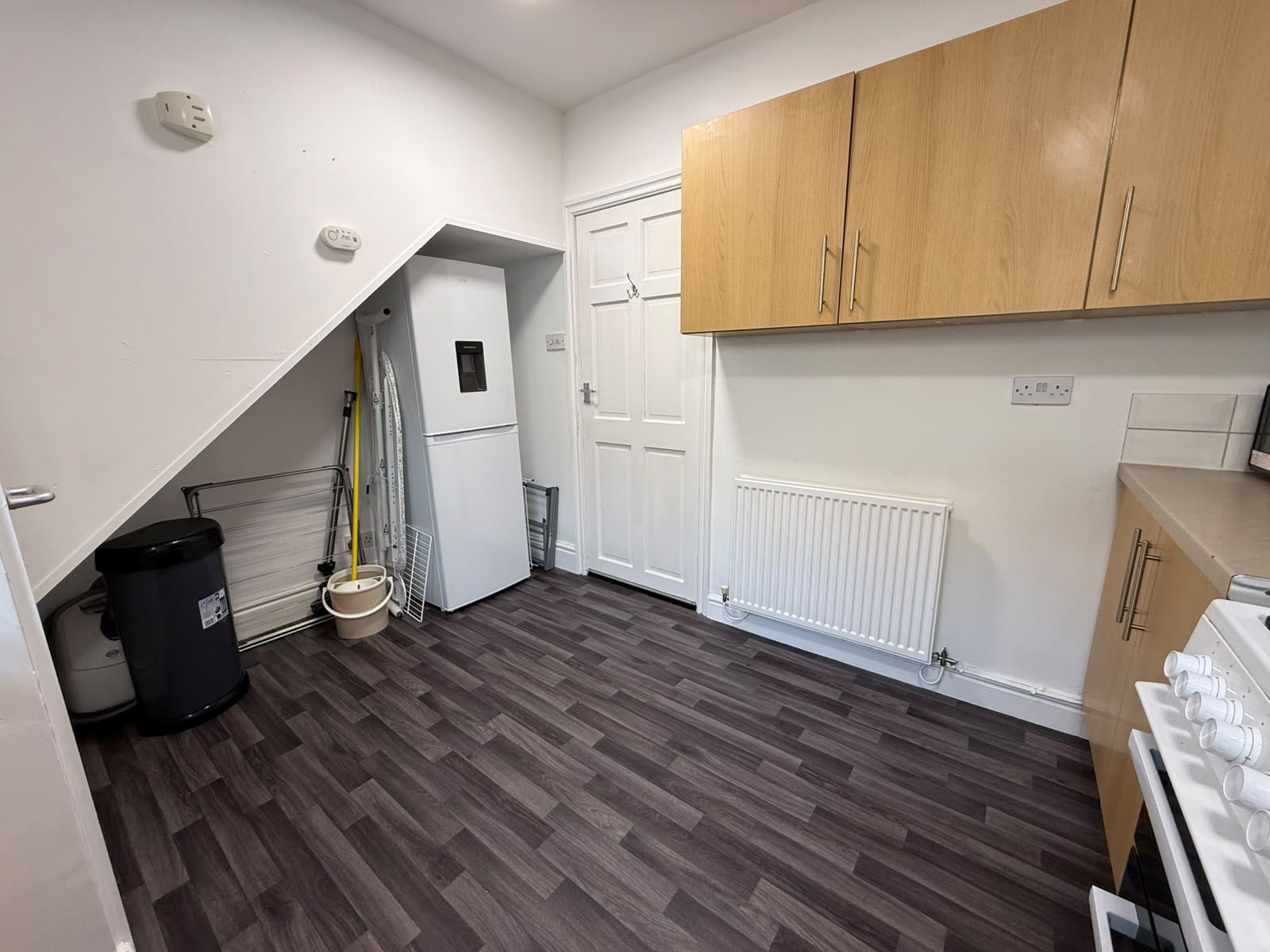 A utility room with a sloped ceiling, white walls, wooden cabinetry, a washing machine, a fridge, and cleaning supplies, with wood-look laminate flooring.